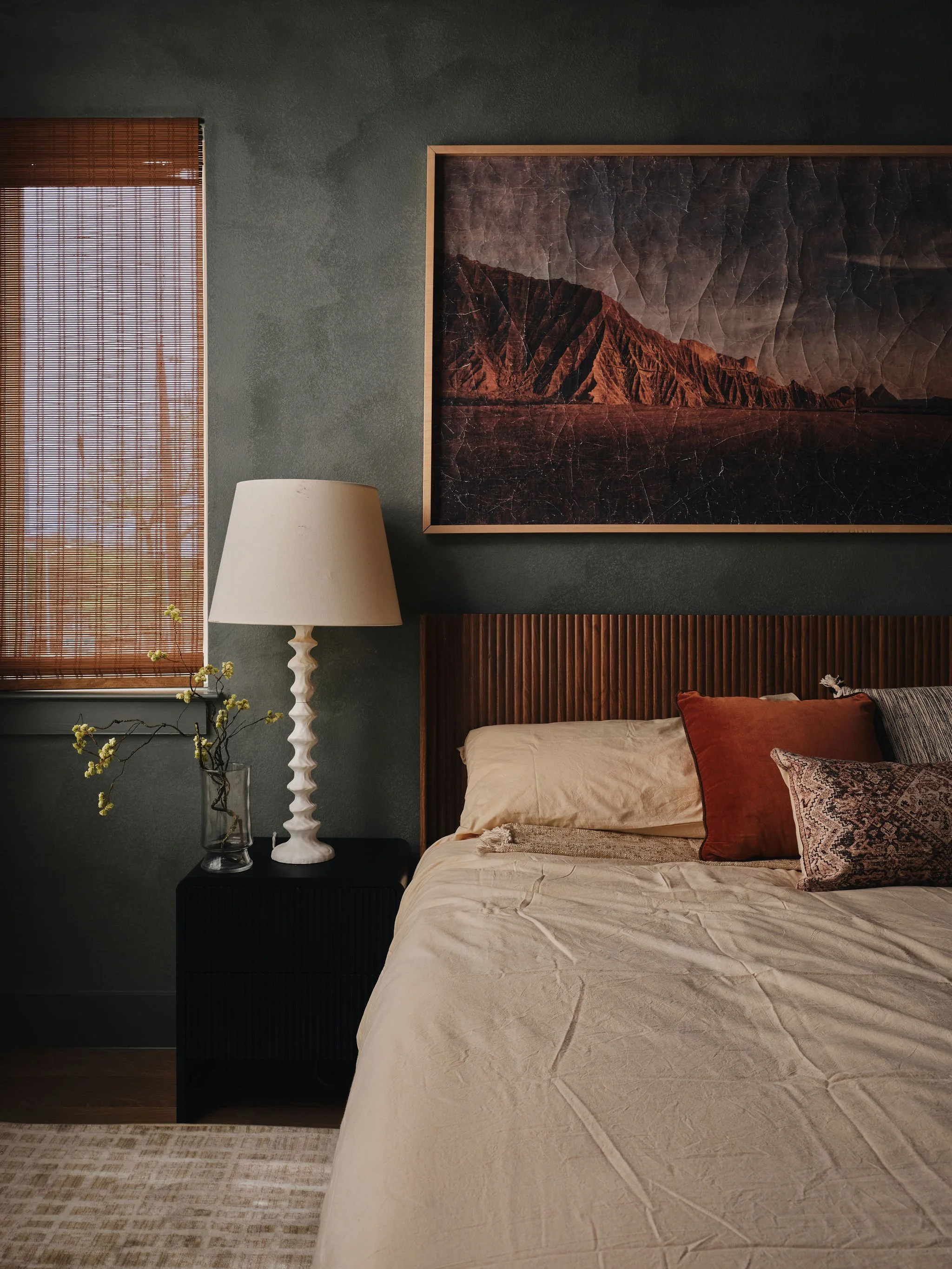 A bedroom with a bed, a nightstand, a lamp, and framed artwork on the wall. The bed has cushions, and there is a window with wooden blinds.