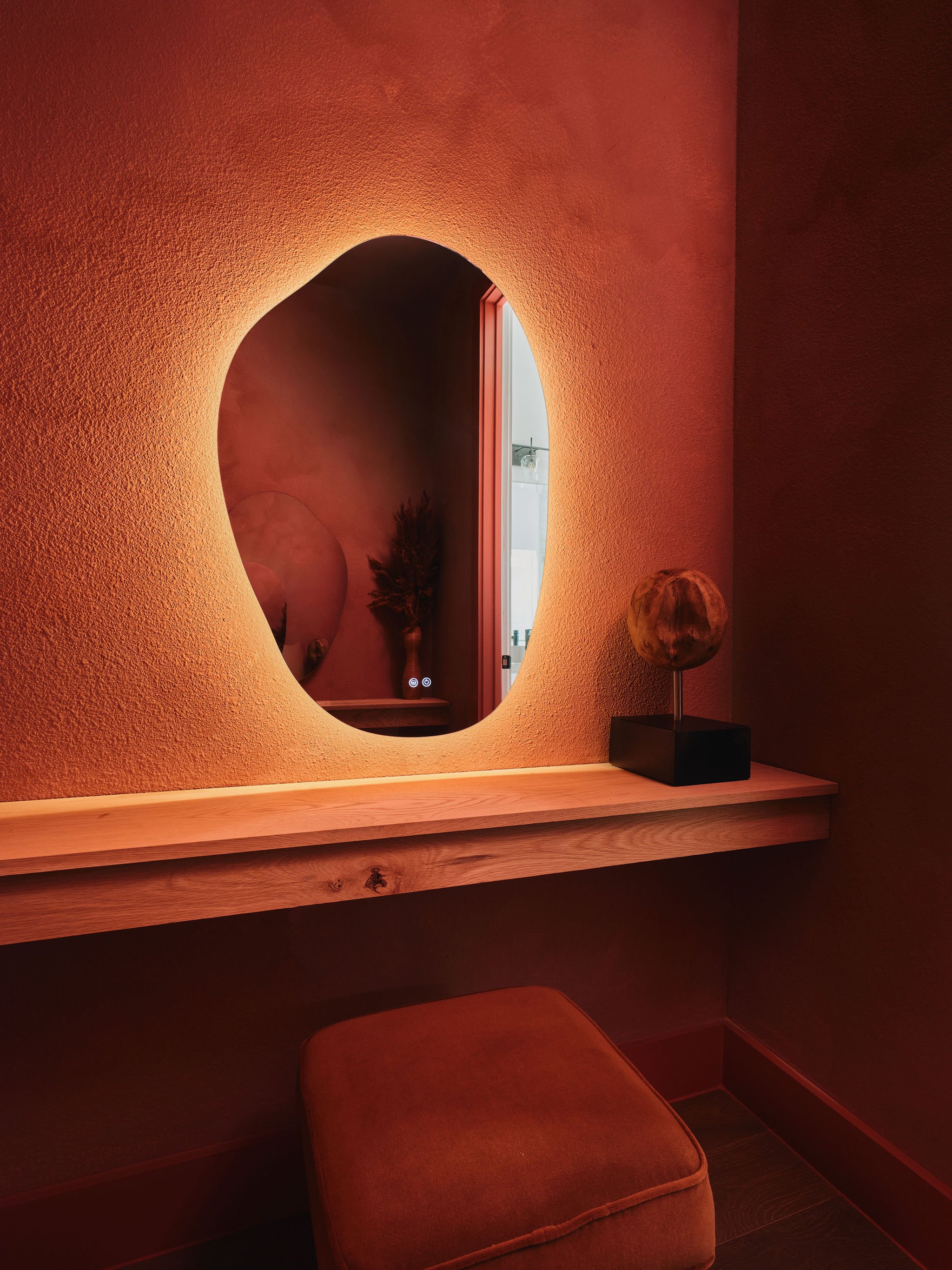 A dimly lit interior with a textured orange wall, a wooden shelf, a mirror, a lamp, and an ottoman.