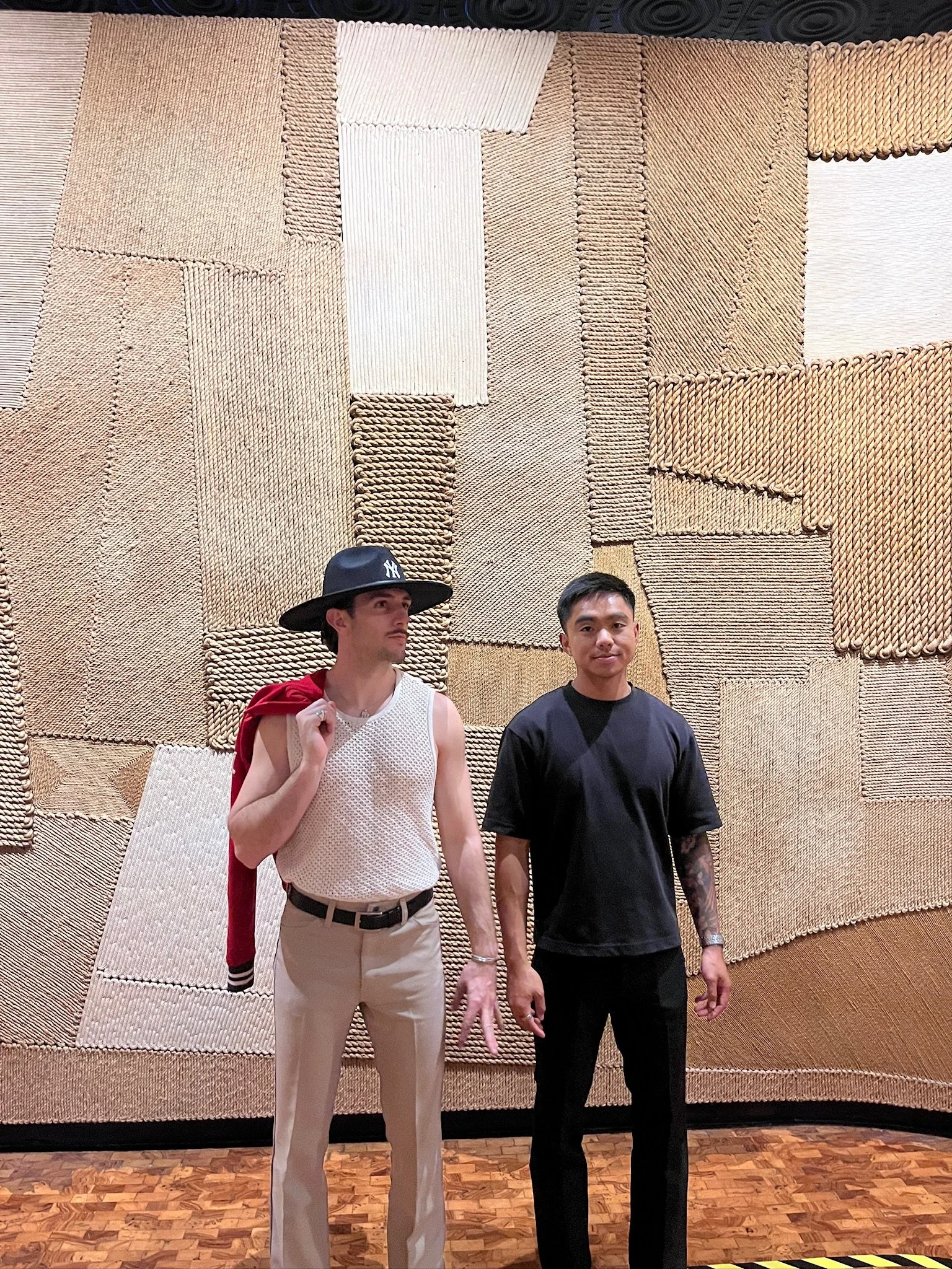 Two men standing in front of a textured wall with woven patterns. One is wearing a white sleeveless shirt, beige pants, and a black hat with a New York Yankees logo, holding a red jacket over his shoulder. The other is wearing a black T-shirt and black pants. The floor is wood, and there is a black and yellow striped line at the bottom edge of the image.