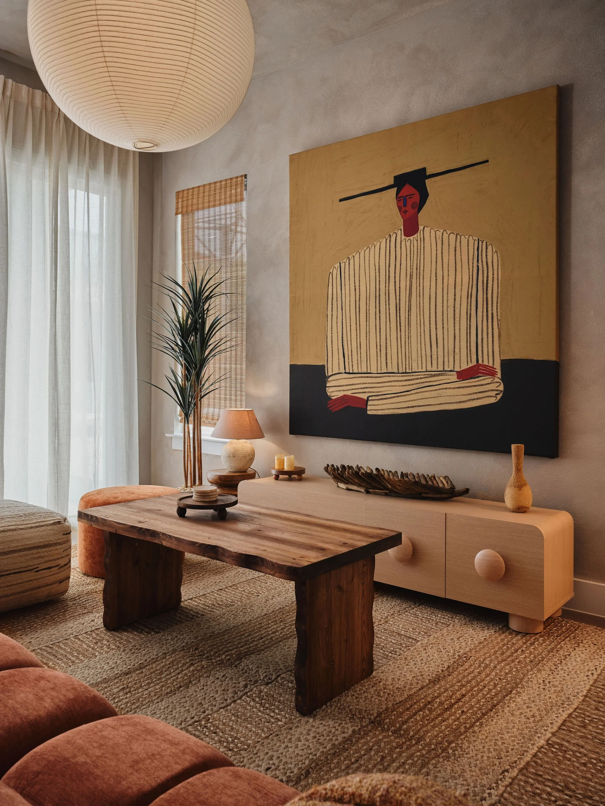 A cozy living room with natural light, featuring a wooden coffee table, a large abstract artwork of a woman with an elongated face and neck hanging on the wall, a tall potted plant, a table lamp, and earthy tones in the furniture and decor.