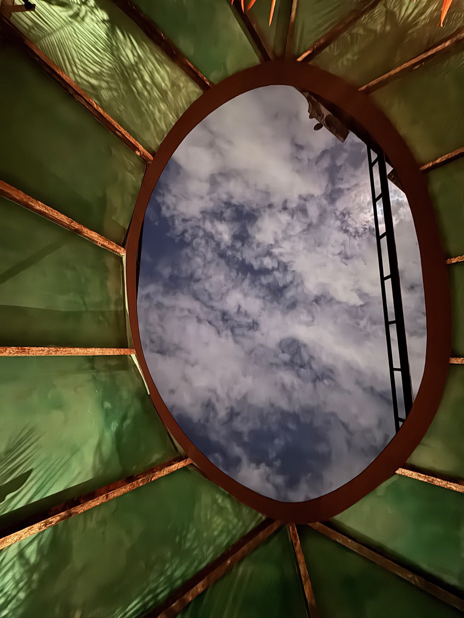 Looking up through a circular opening in a green thatched roof, revealing a partly cloudy sky with some clouds and sunlight.