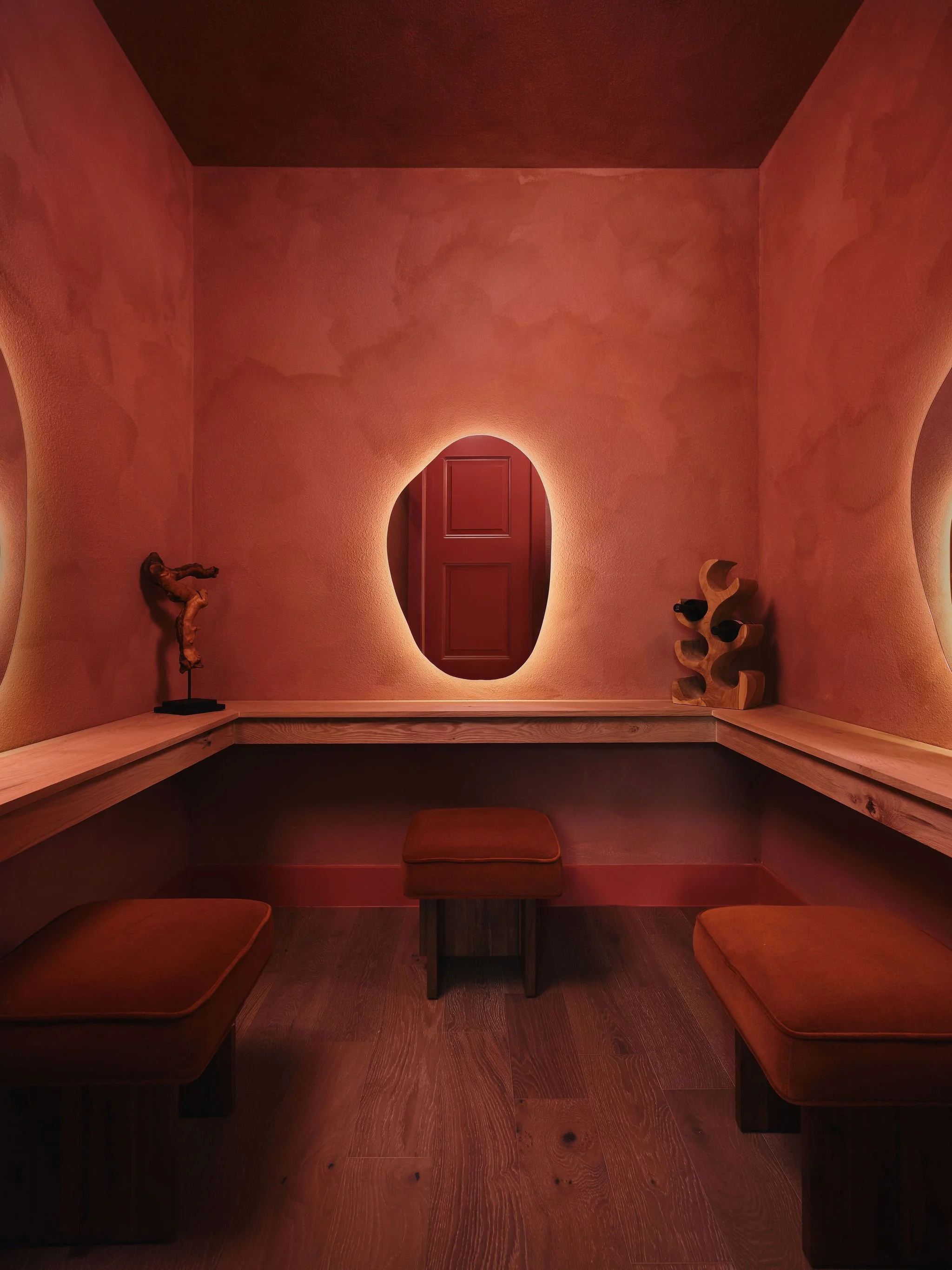 A cozy, dimly lit nook with curved, pinkish-orange walls and built-in wooden seating cushions with matching pillows. A small wooden table is in the center, flanked by three seating cushions on each side. A wooden sculpture of a human figure is on the