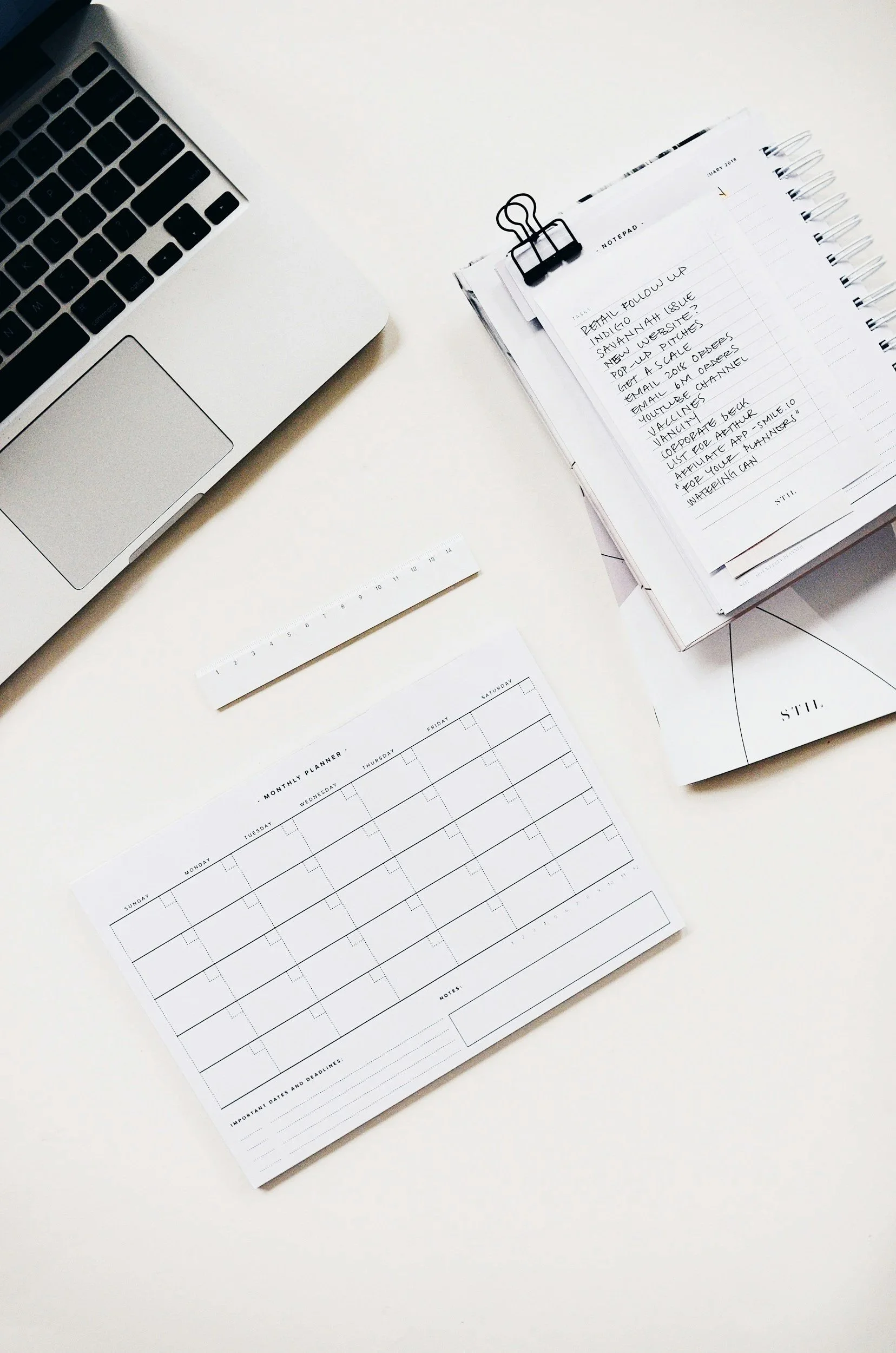 White desk with a laptop, calendar, planner, and note pages, some held with a black binder clip.