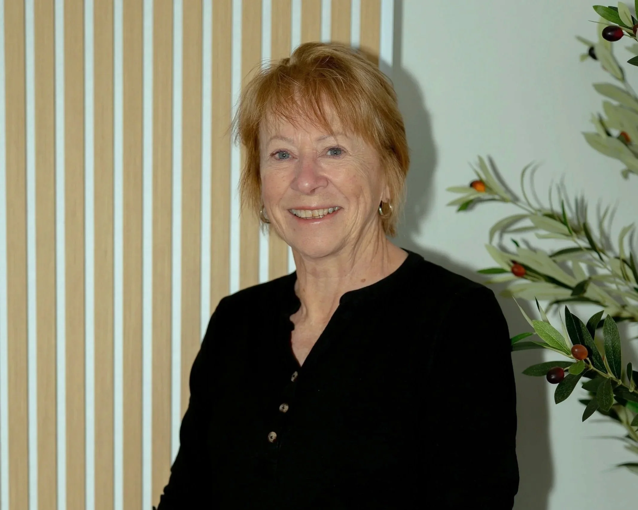 A woman with short red hair, wearing a black top with buttons, smiling indoors near a wooden slat wall and an olive tree with small dark olives.