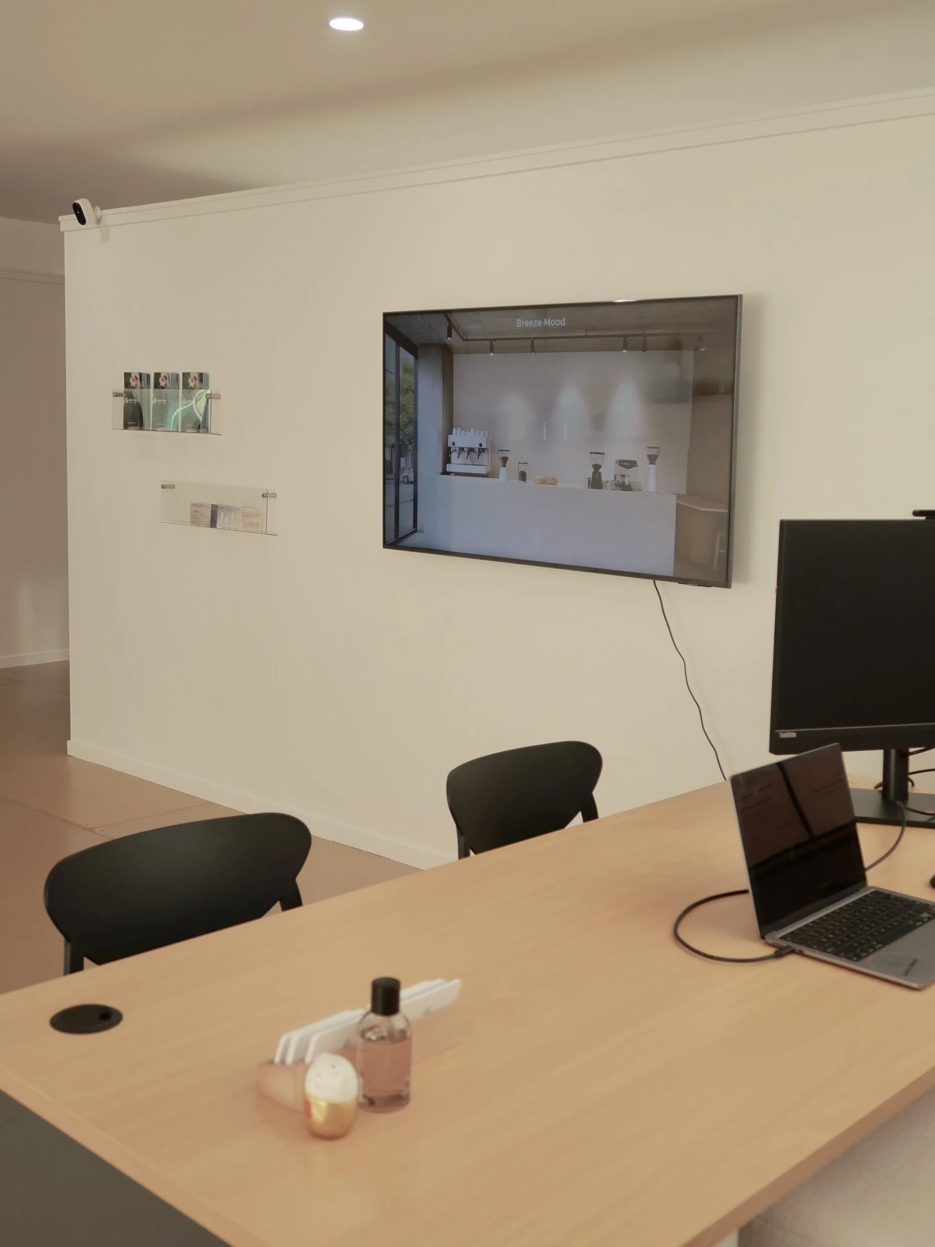 Office conference room with a large wooden table, black chairs, a large screen on the wall displaying a coffee shop, a smaller monitor, and laptop on the table. There are some bottles and small containers on the table.