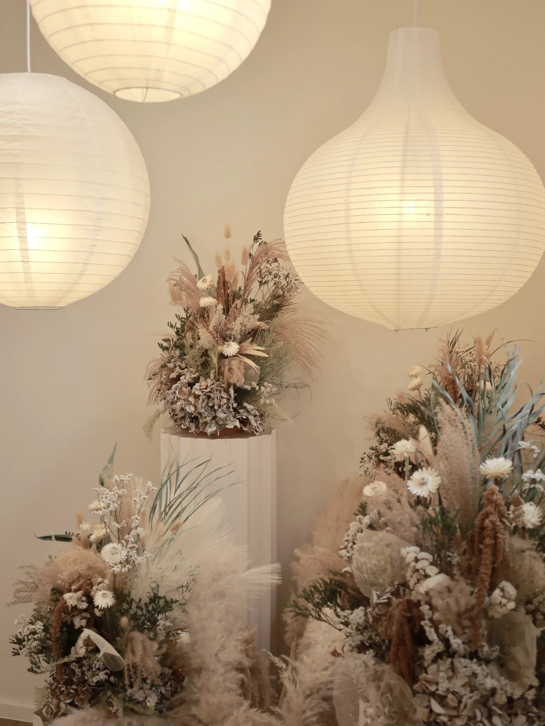 Arrangement of cream and beige dried flowers with pampas grass under three hanging white paper lanterns.