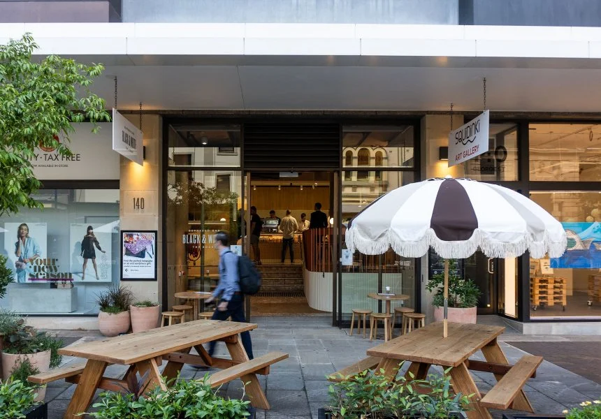 Street view of an art gallery entrance with wooden picnic tables and a striped umbrella outside, plants and storefronts nearby.