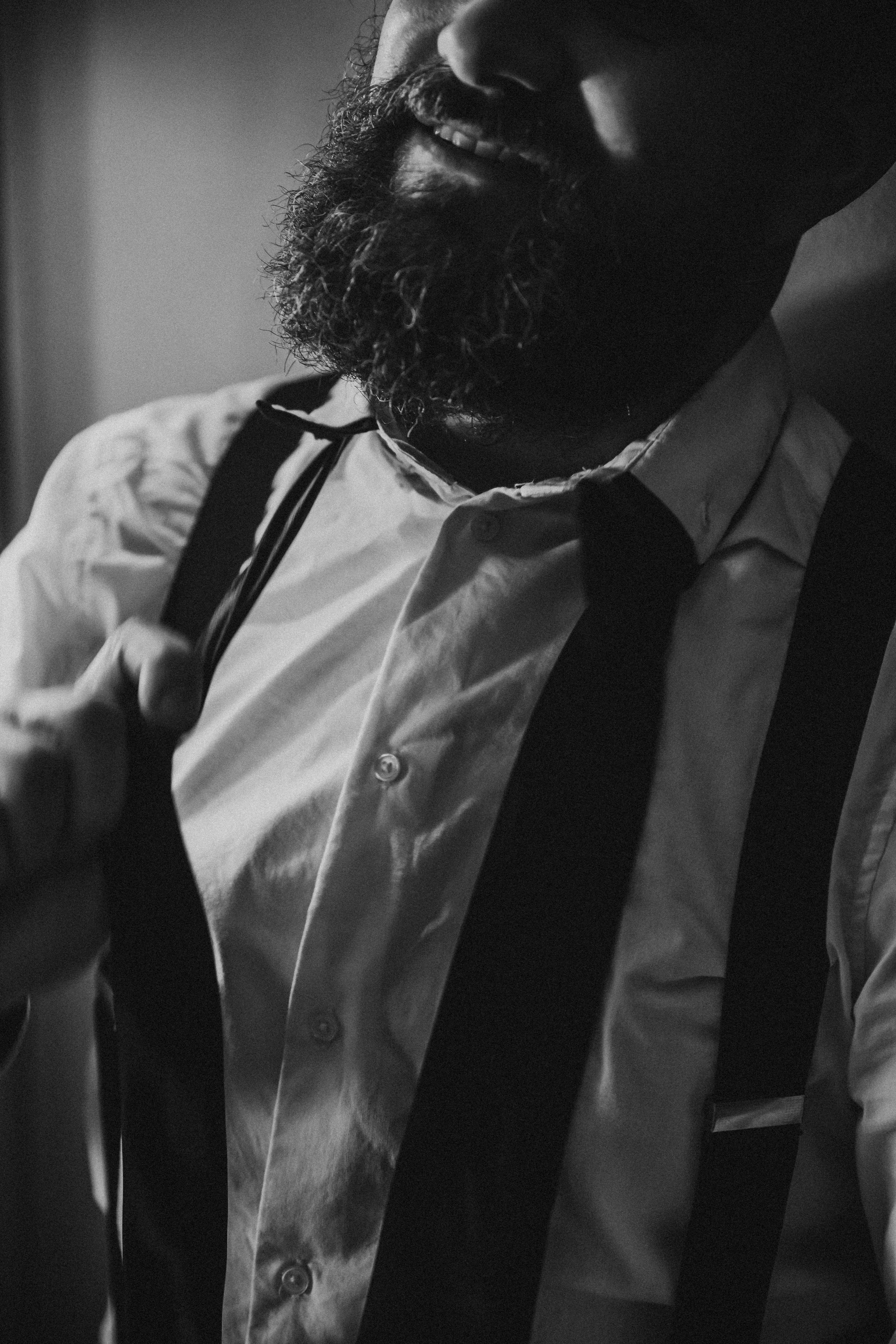 Close-up of a man with a beard, wearing a white dress shirt, black suspenders, and a black tie, smiling and adjusting his suspenders.
