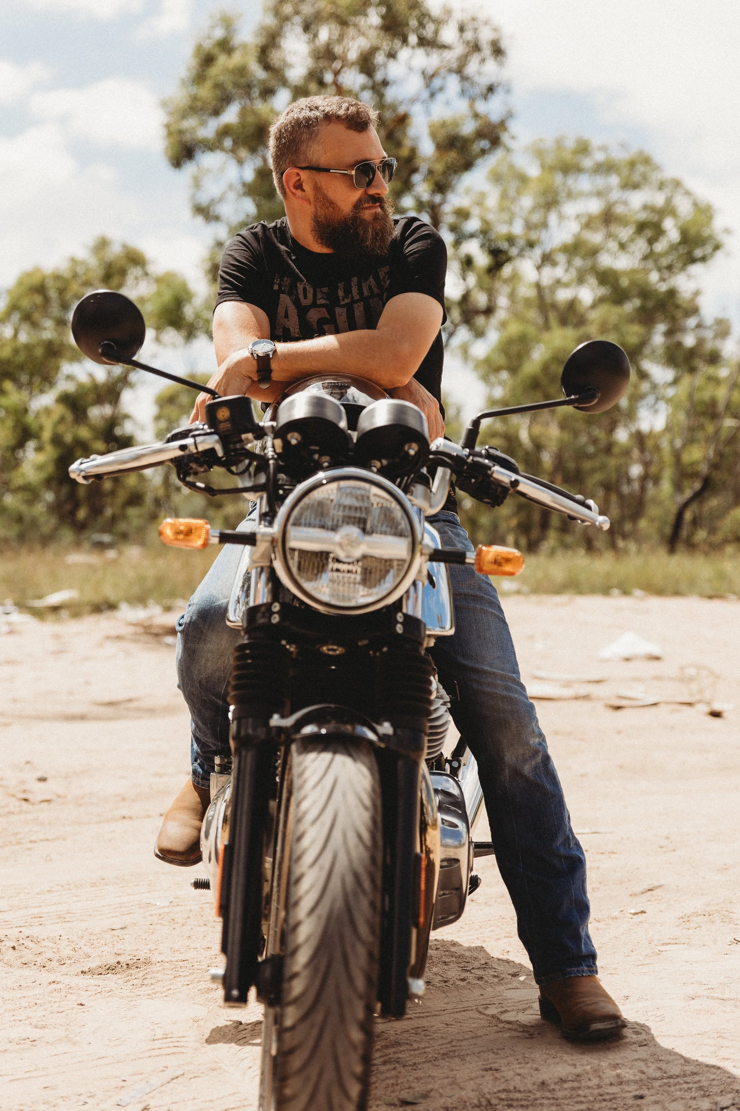 A man with sunglasses and a beard is sitting on a motorcycle, wearing a black T-shirt and jeans, in an outdoor setting with green trees and a dirt ground.