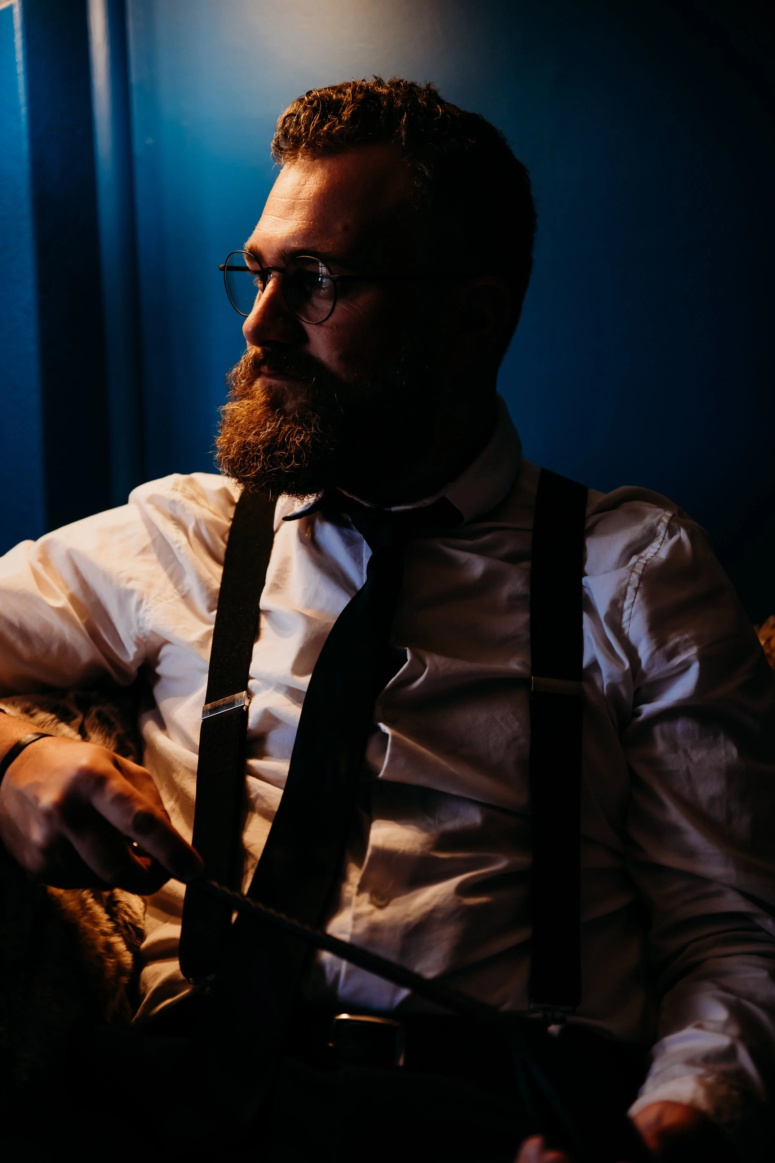 A bearded man wearing glasses, a white shirt, a black tie, and suspenders, sitting with a cat on his lap, in a dimly lit setting with a blue background.
