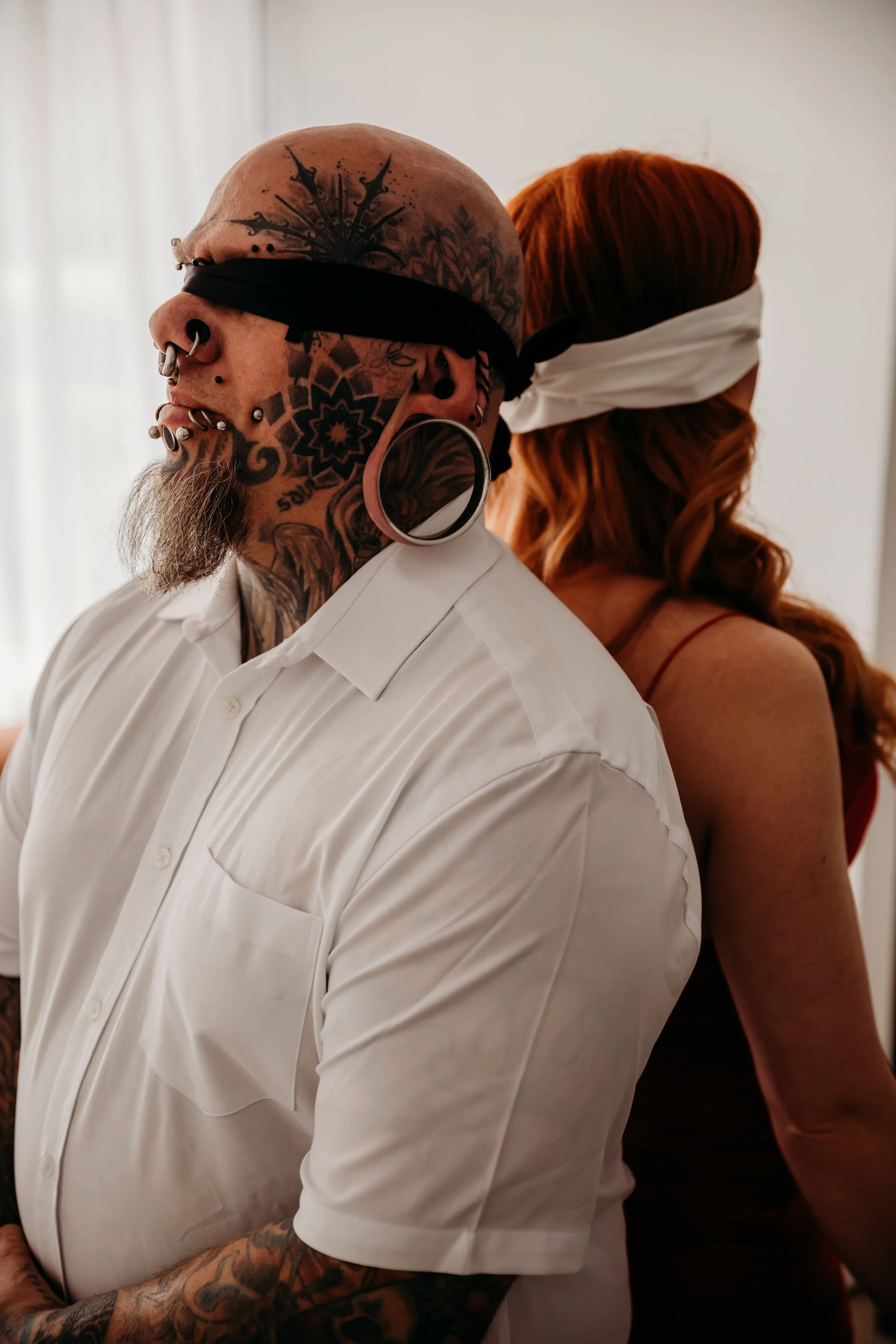 A tattooed man with piercings and large gauge earrings is standing with a woman who has red hair and is wearing a white bandana over her eyes. Both are facing sideways.