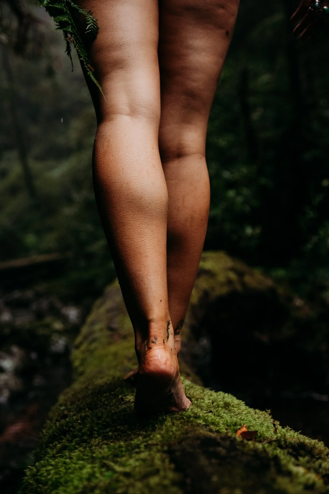 A close-up of a person's bare legs and feet walking on a moss-covered log in a forest setting.