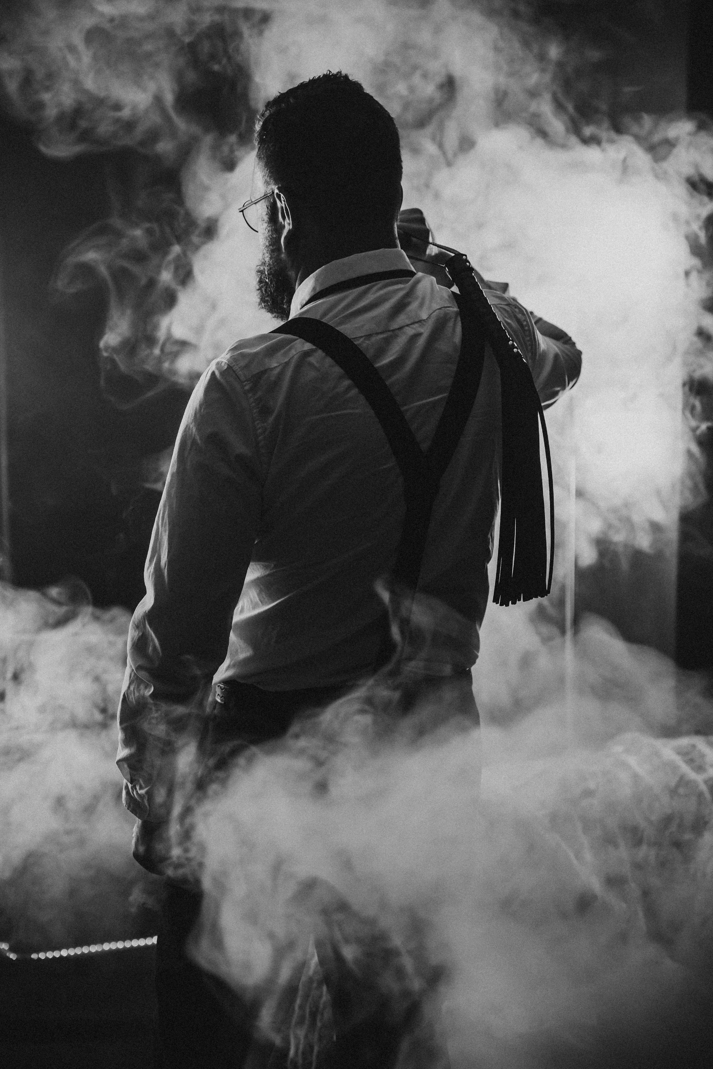 A man with a beard and glasses standing in a smoky environment, adjusting a mask or respirator on his face, backlit with smoke or vapor around him.