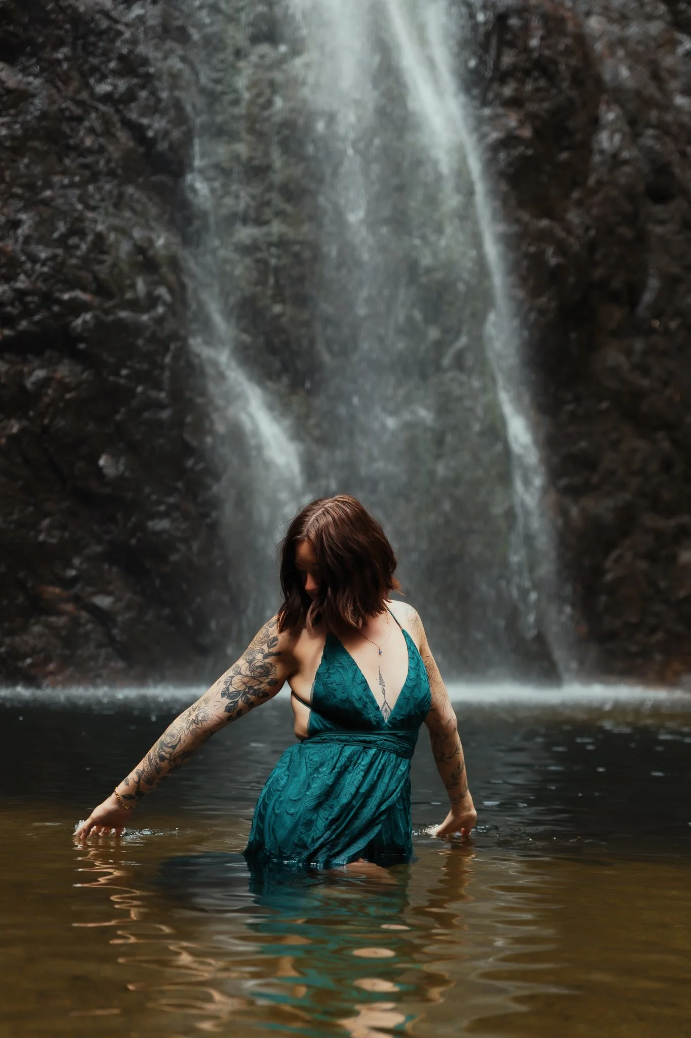A woman with tattoos on her arms standing in a body of water in front of a waterfall, wearing a teal dress.