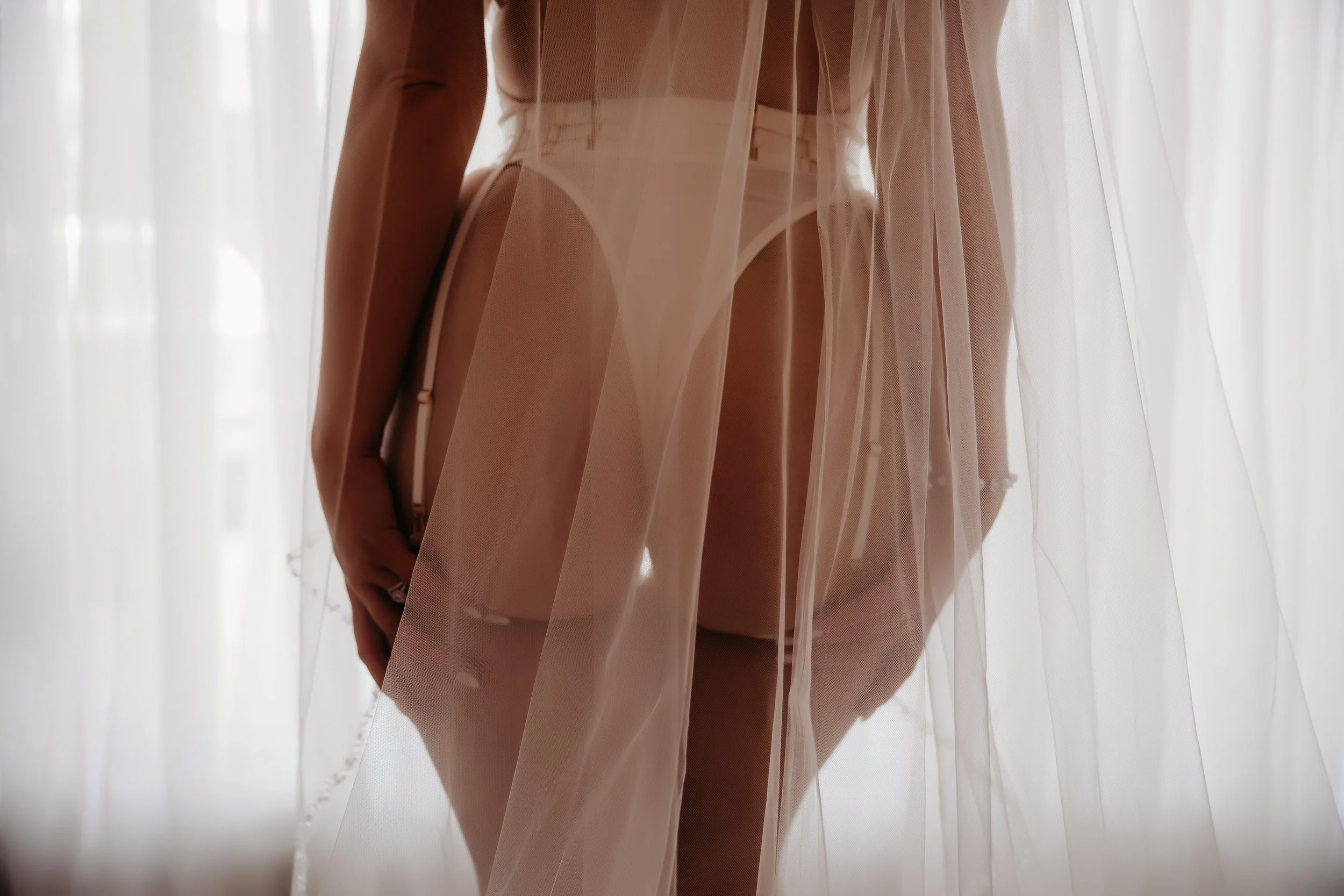 A person dressed in nude-colored lingerie and a sheer, flowing robe standing behind semi-transparent curtains, with light filtering through.