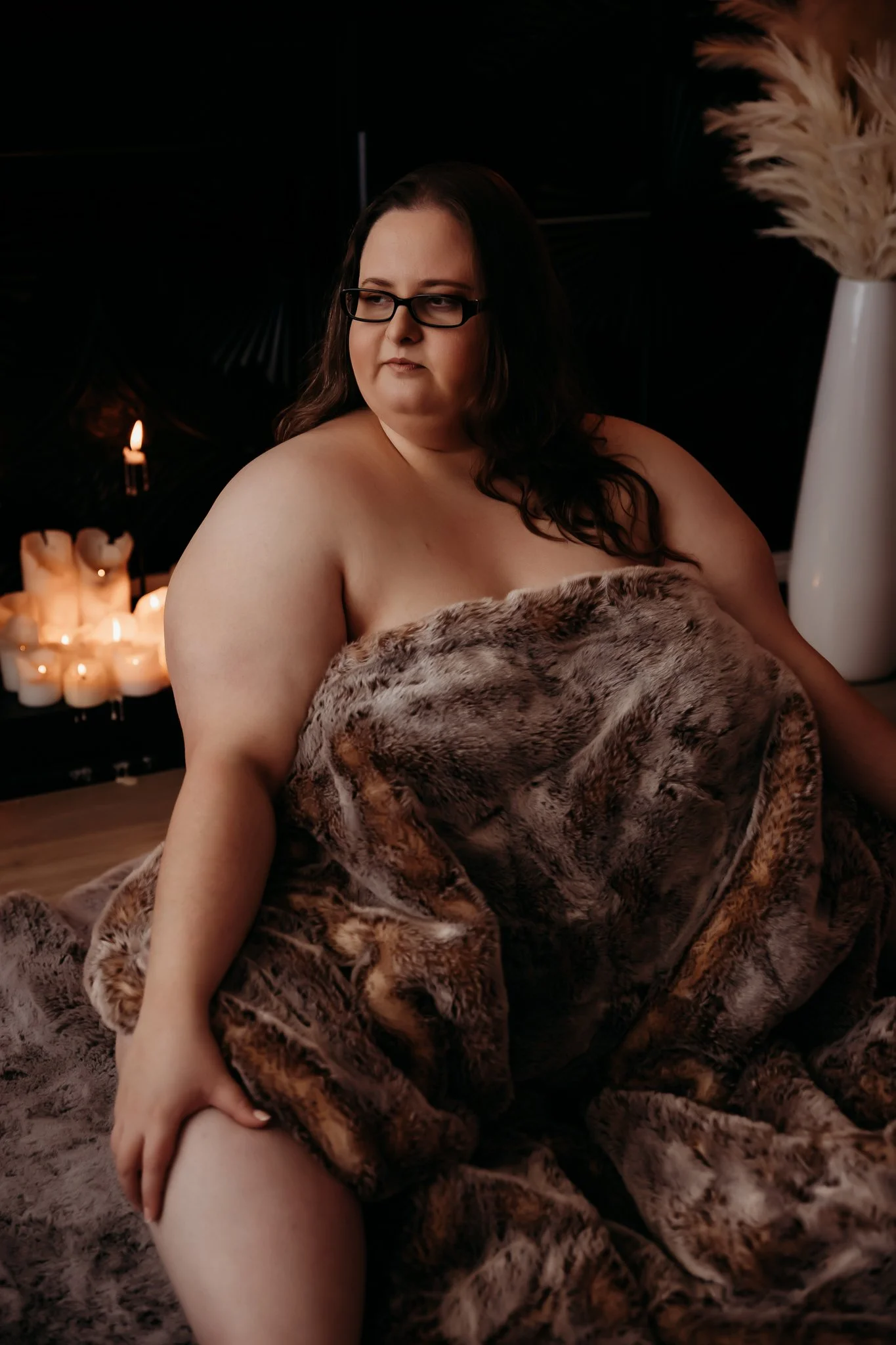 A woman with glasses and long dark hair sitting on a furry blanket draped over her body in a dimly lit room, with candles in the background.