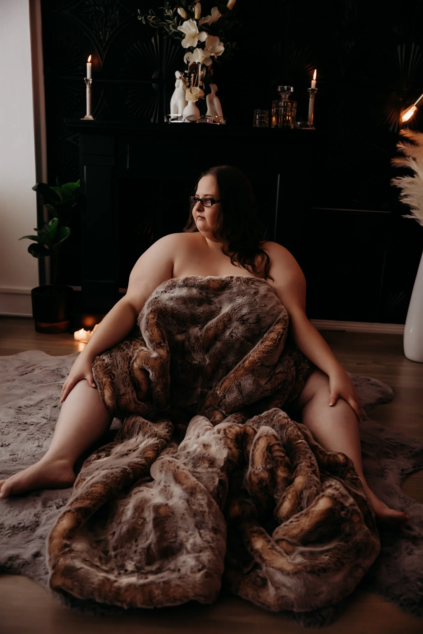 A woman sitting on the floor wrapped in a fur blanket, with a dark background featuring candles, vases, and plants, creating a warm, intimate setting.