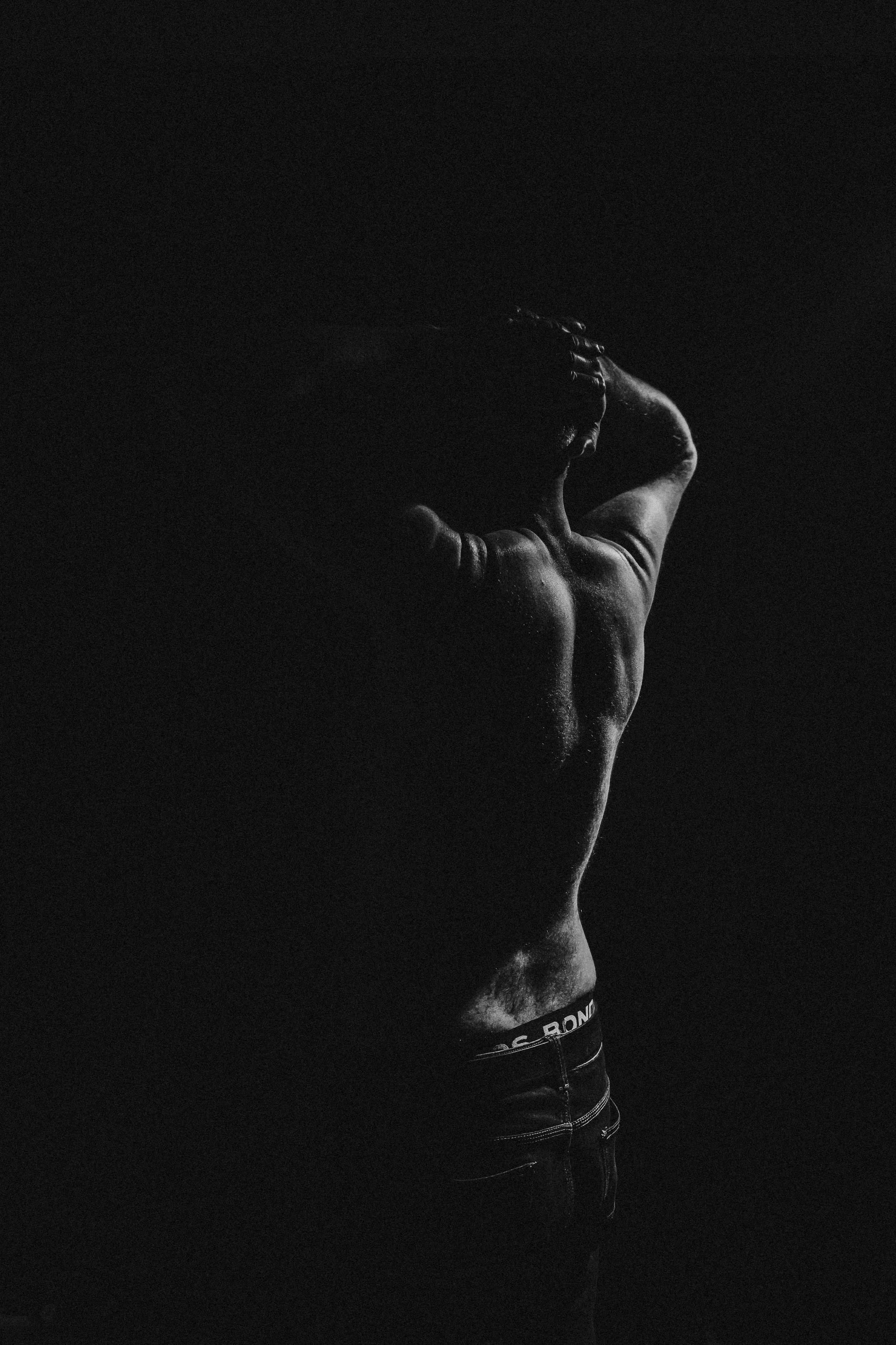 A black and white photograph of a shirtless man posing with one arm raised behind his head, showcasing his muscular back and side profile.