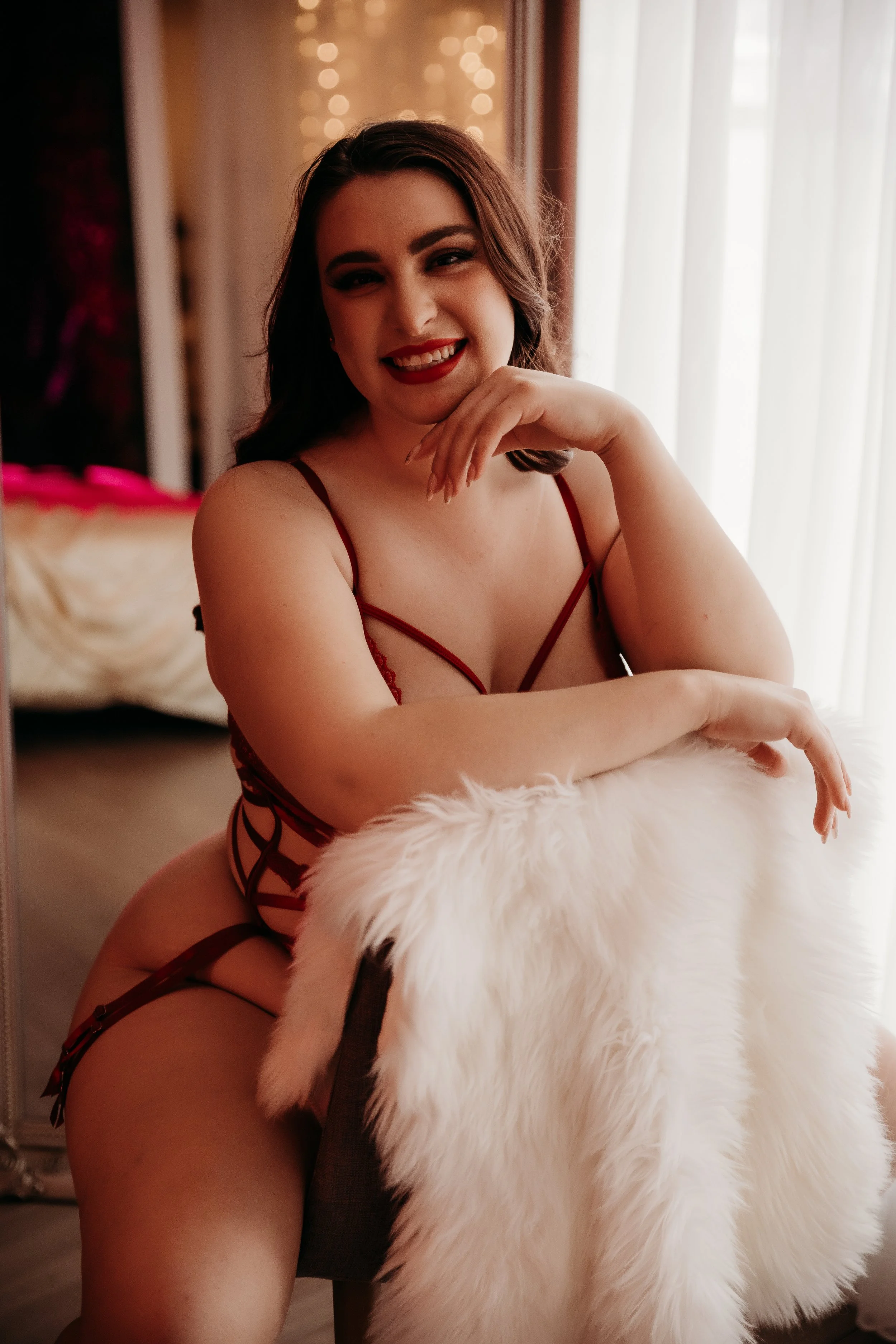 A woman with dark hair, wearing red lingerie, smiling and sitting with her elbow resting on a fluffy white fur object, in a warmly lit room with sunlight and string lights in the background.