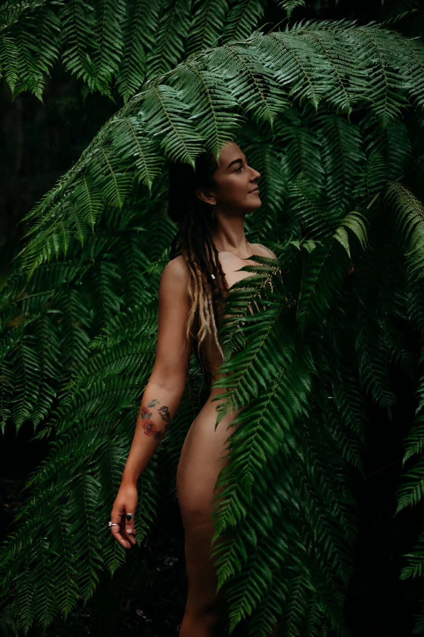 A nude woman with tattoos on her arm and dreadlocks in a lush green forest, partially obscured by large fern leaves.