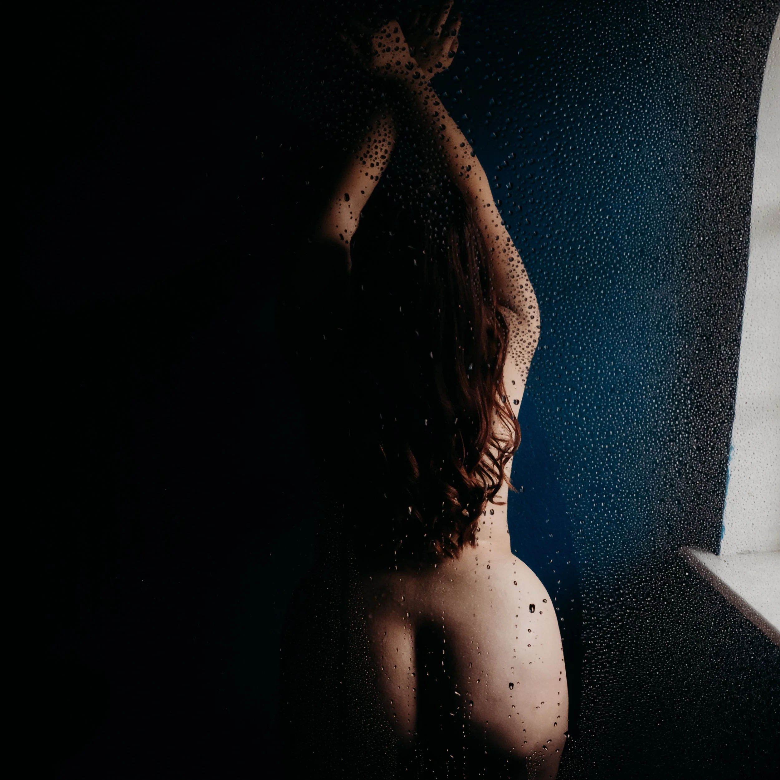 Nude woman with long hair standing near a window with rain droplets on the glass.