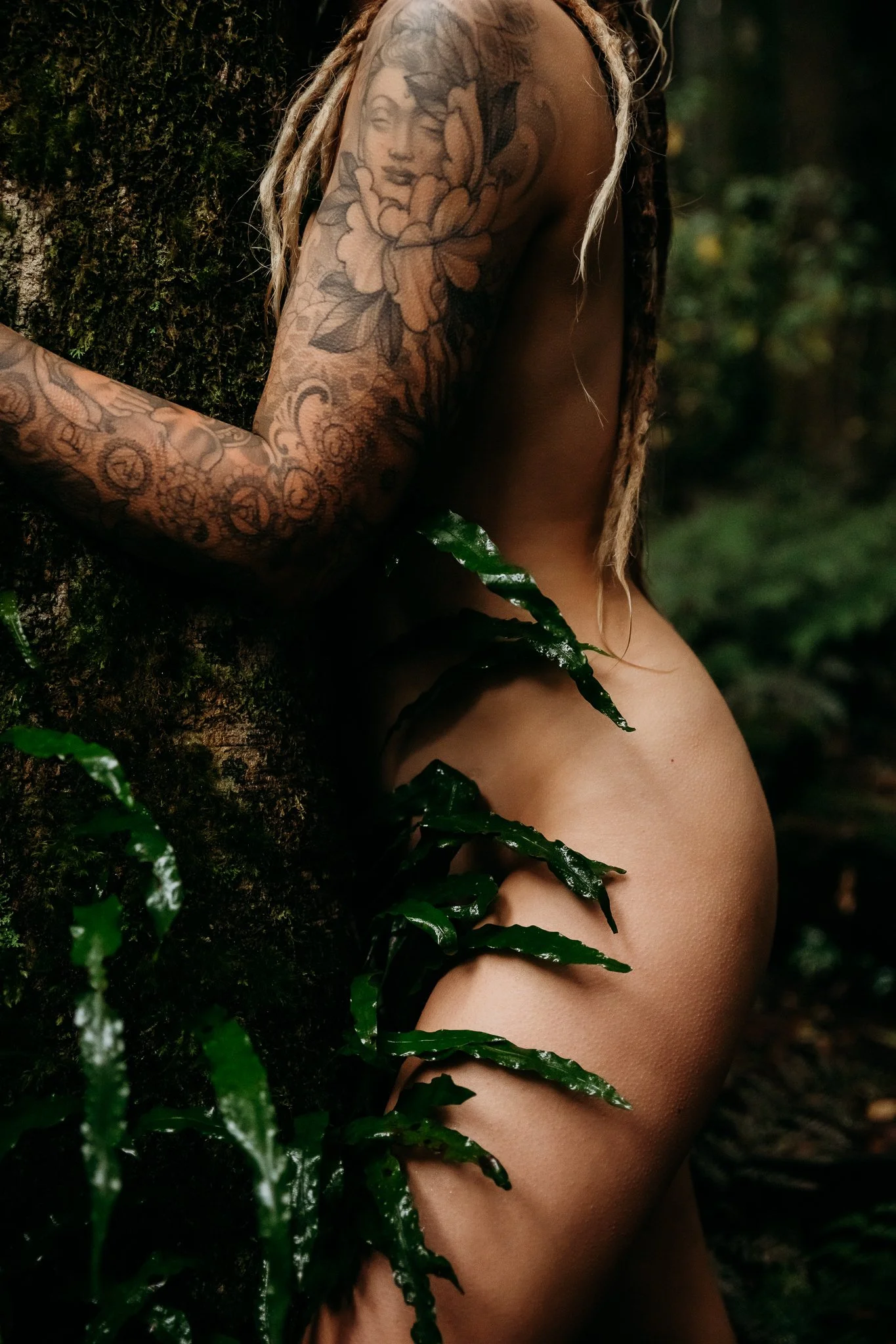 Close-up of a woman with tattoos on her right arm, standing outdoors in a forest, partially covered by green leaves.