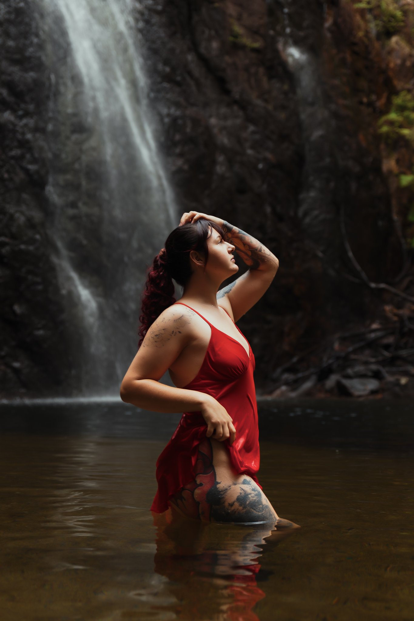 A woman with tattoos in a red dress standing knee-deep in water near a waterfall in a forest.