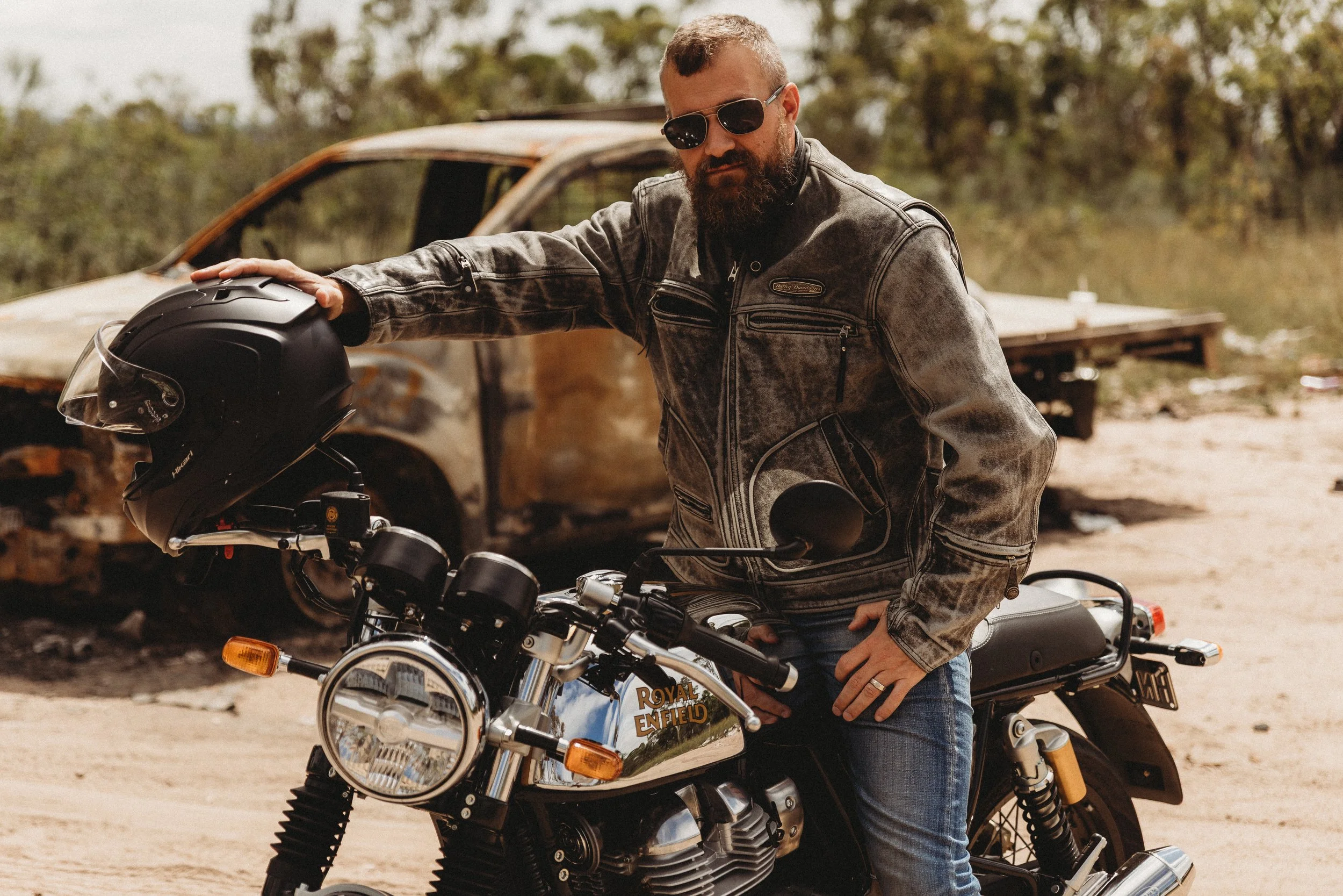A bearded man wearing sunglasses and a black leather jacket, standing next to a black Royal Enfield motorcycle, holding a black helmet. Behind him is a rusty, abandoned car in a desert-like environment.