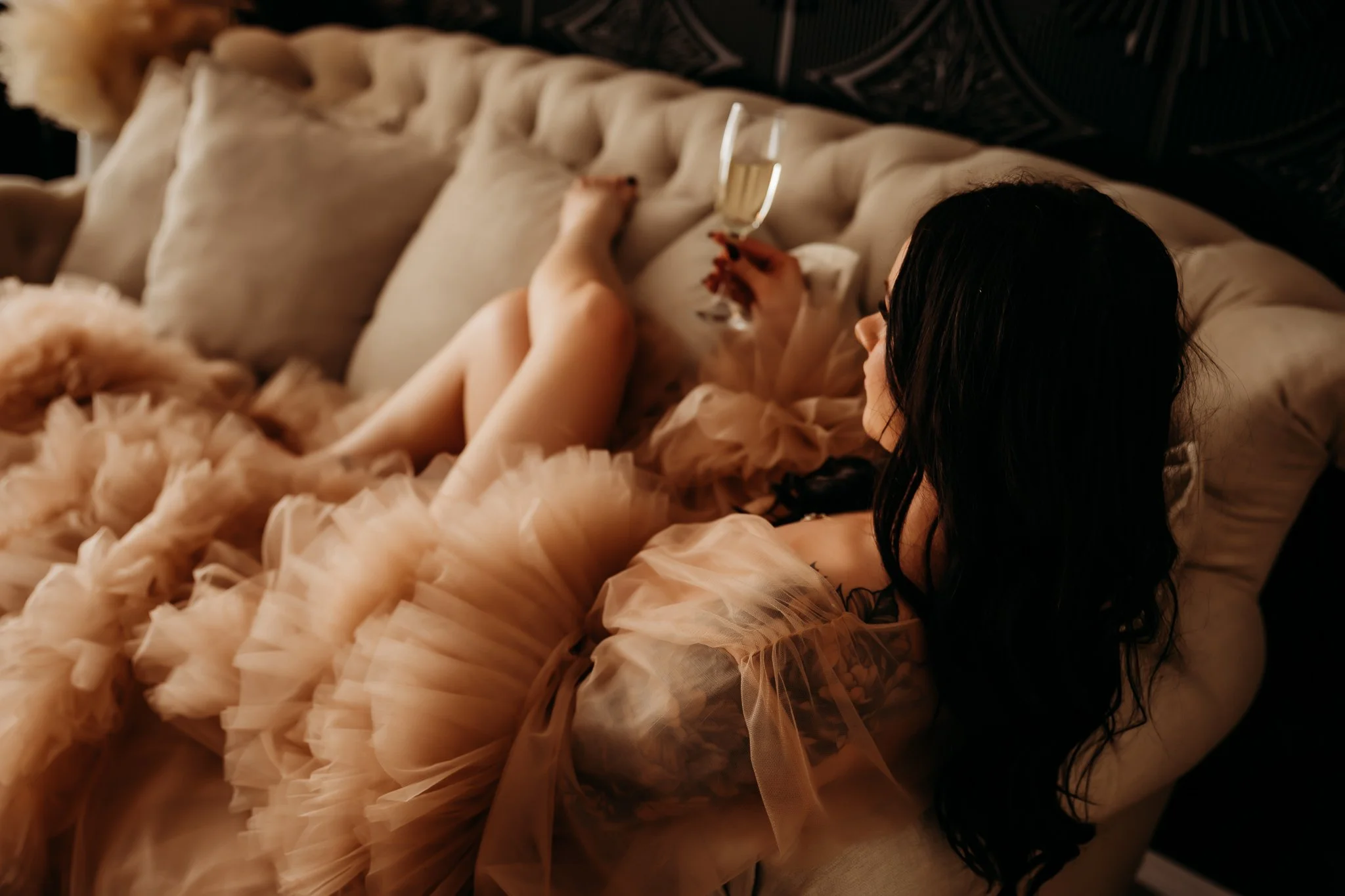 Woman with long dark hair wearing a peach-colored, ruffled gown, lying on a plush beige couch, holding a glass of champagne.