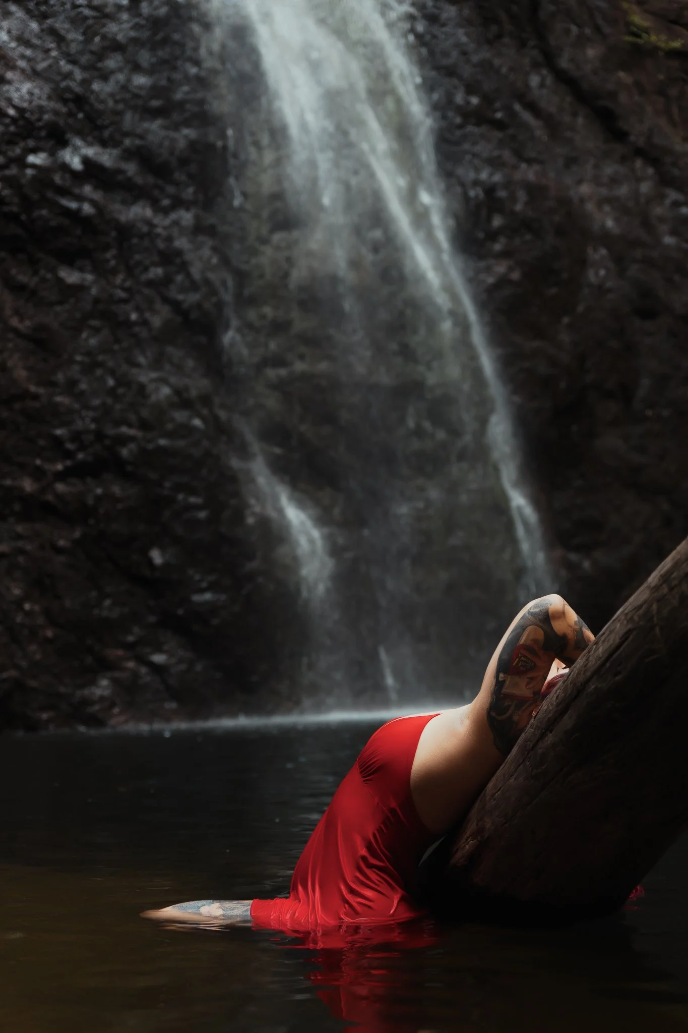 Person in a red swimsuit lying on a log in a body of water, with a waterfall in the background.
