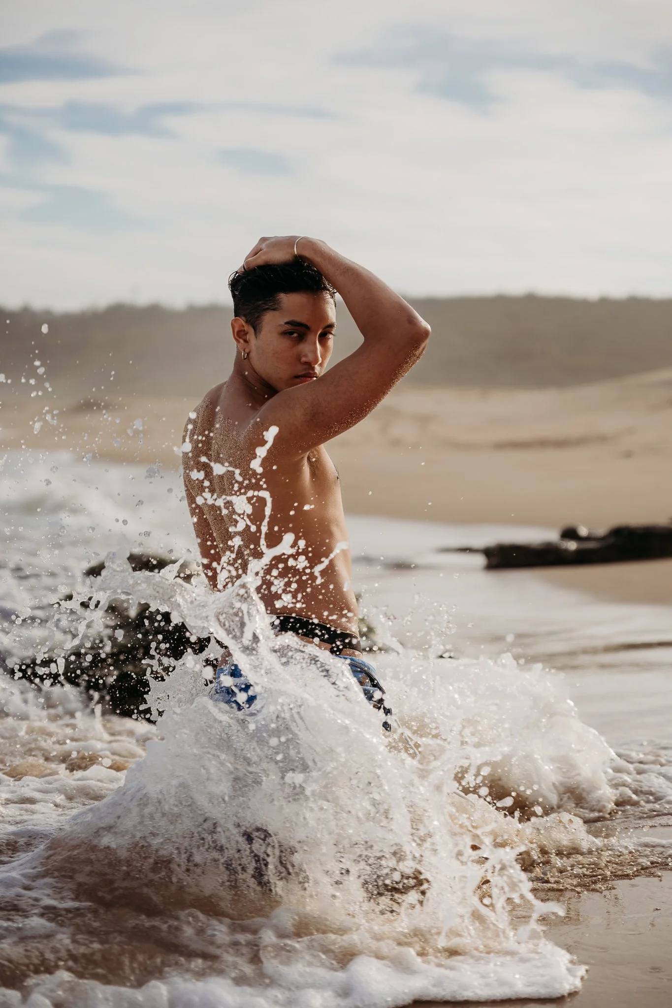 A young man is in the ocean, half-submerged in water, with waves splashing around him. He has dark hair, wears earrings, and is holding his hair back with one hand. The background shows a sandy beach and a cloudy sky.
