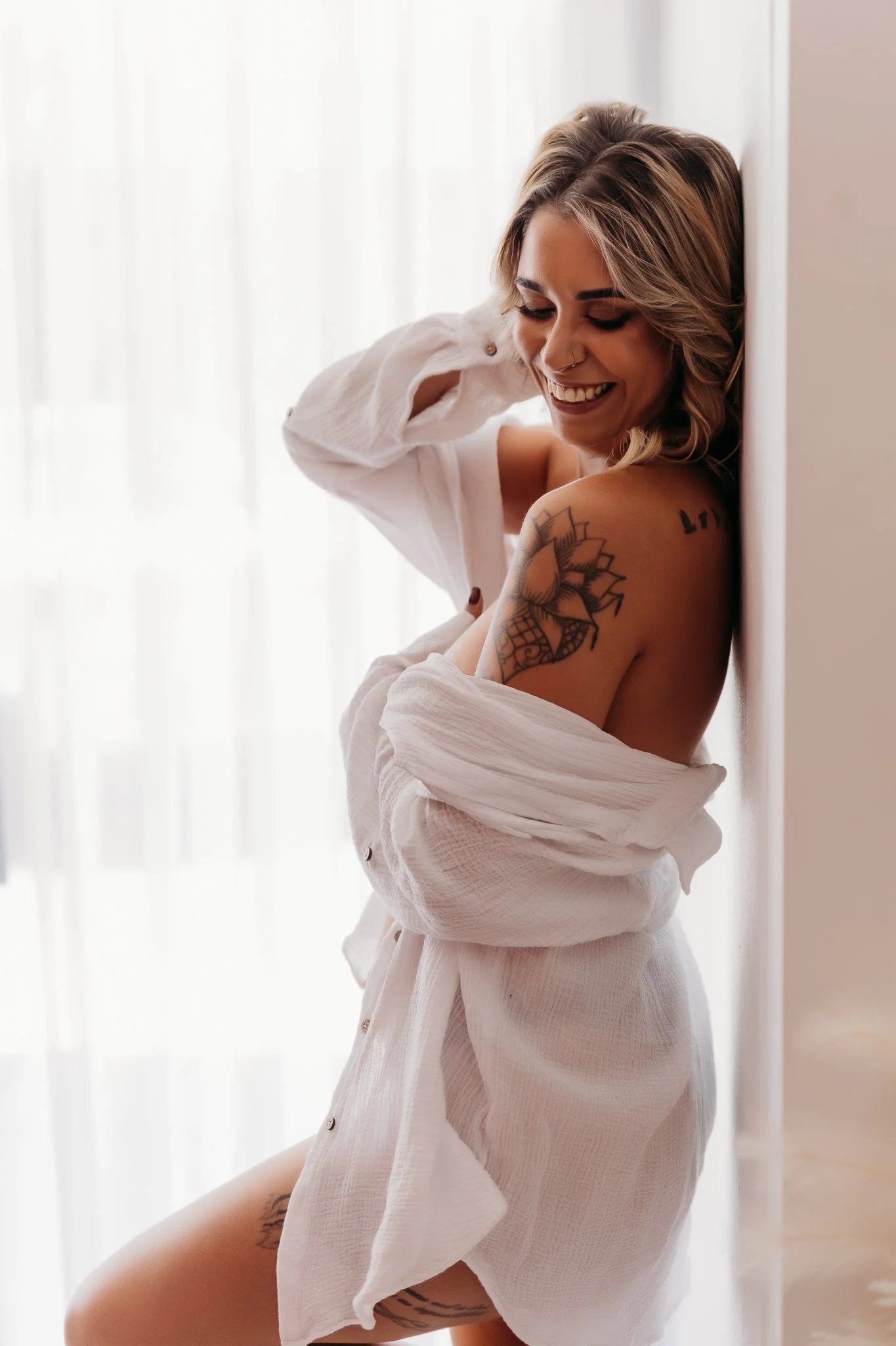 A woman with short blonde hair and tattoos on her arm and thigh, wearing an open white shirt, smiling and leaning against a wall near a window with sheer curtains.
