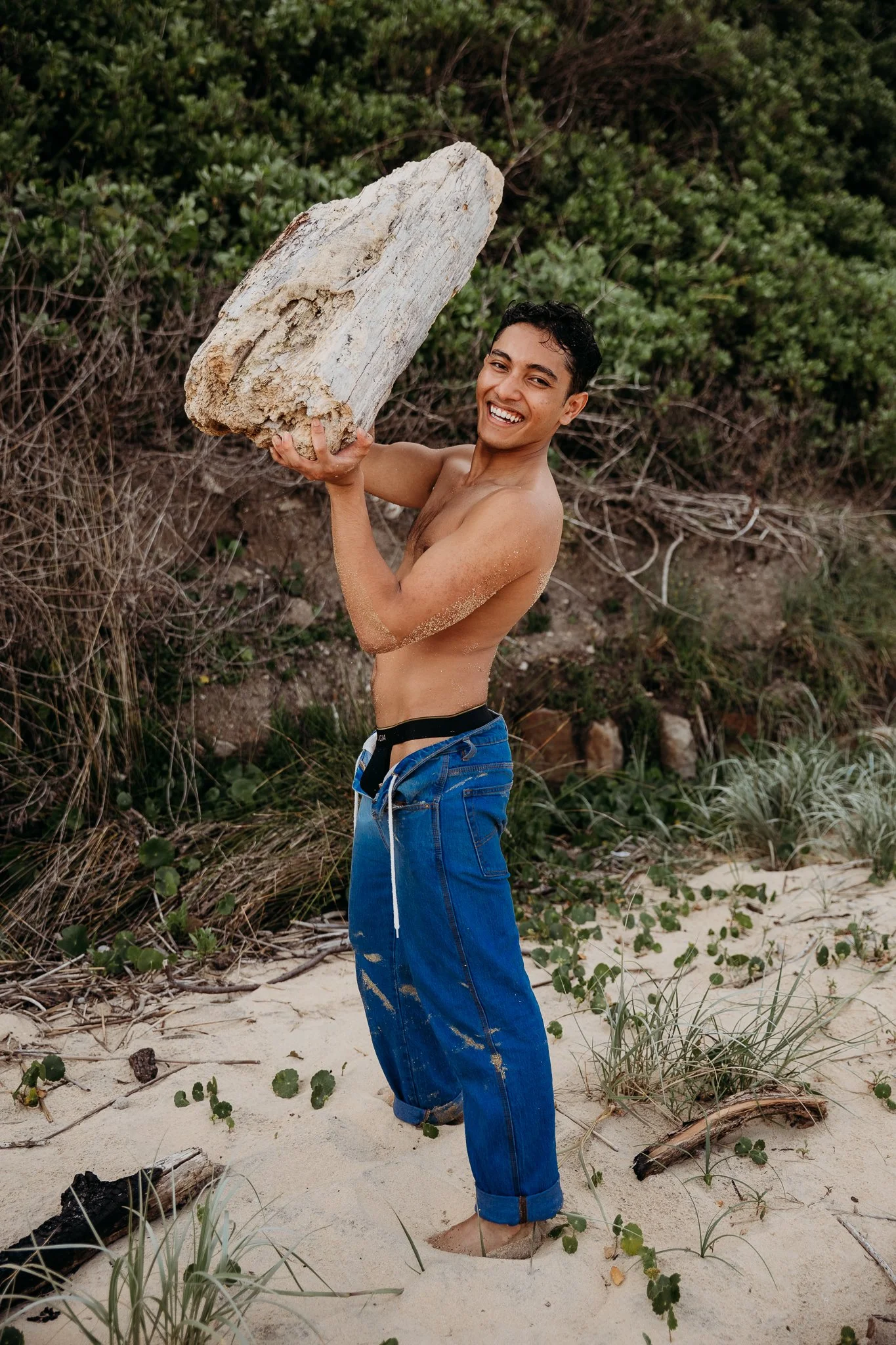 A smiling young man holding a large piece of driftwood on a sandy beach with green vegetation in the background.