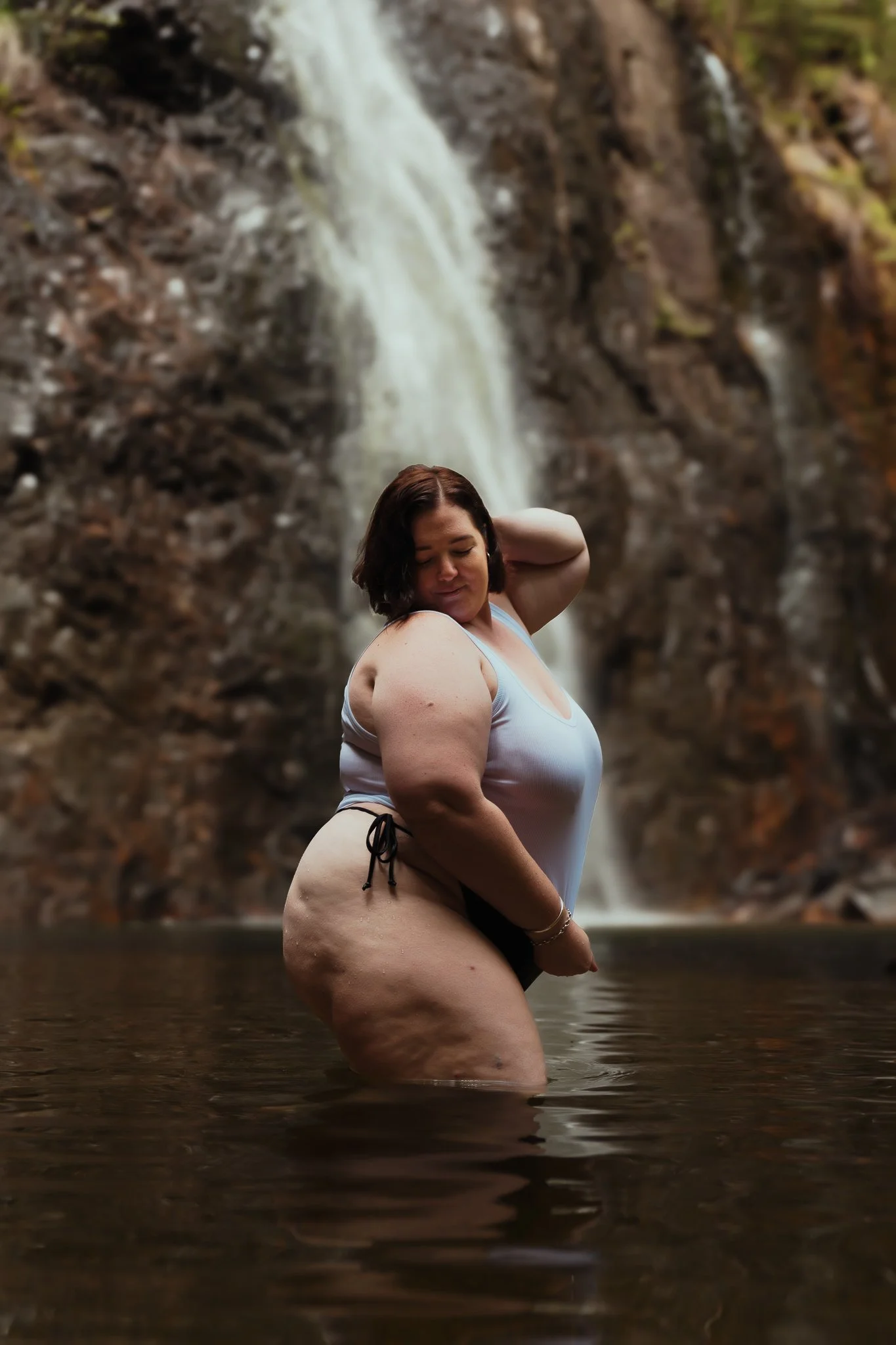 A woman standing in water near a waterfall, wearing a white tank top and black shorts, with her eyes closed and one hand behind her head.