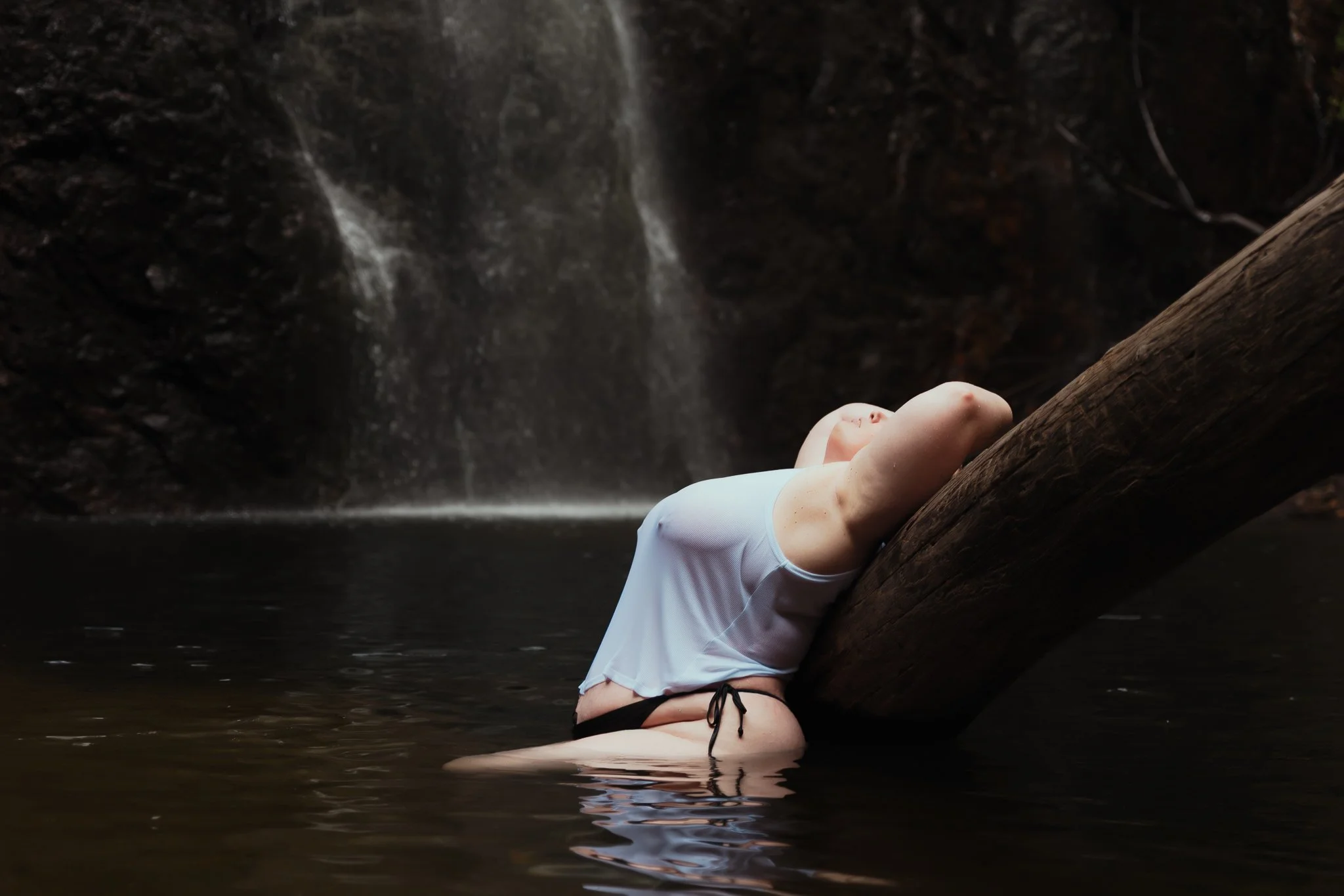 Person in a white top and black bottom leaning back against a large log in a body of water near a waterfall.