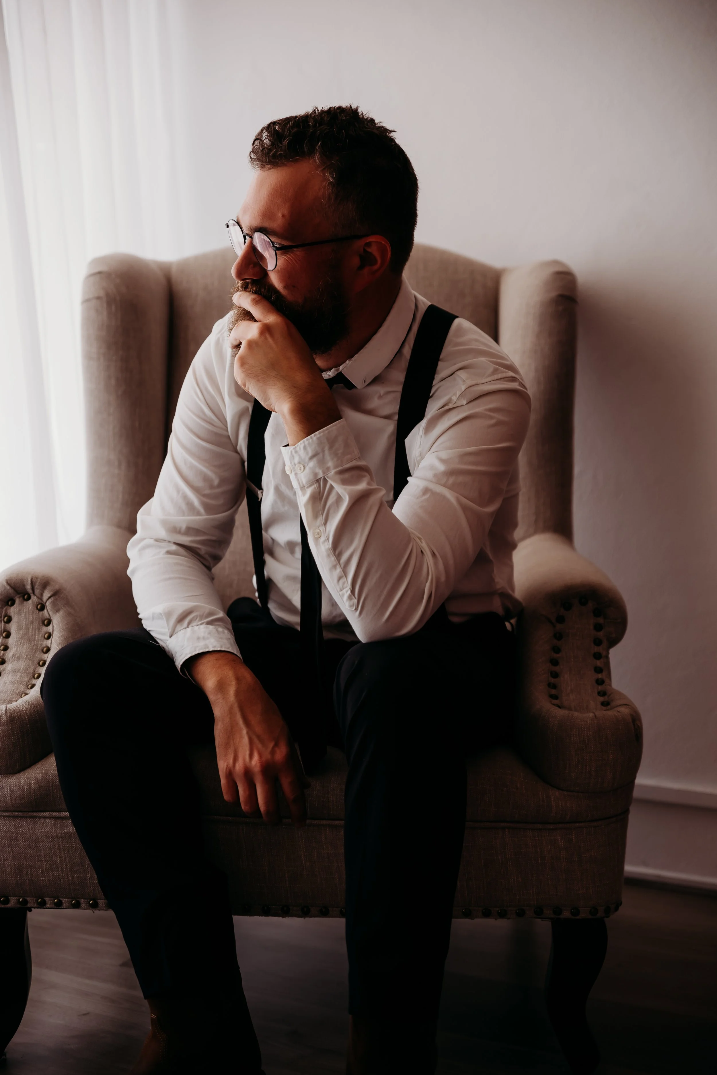 A man with glasses, a beard, wearing a white shirt and black suspenders, sitting on a beige armchair and looking thoughtful.