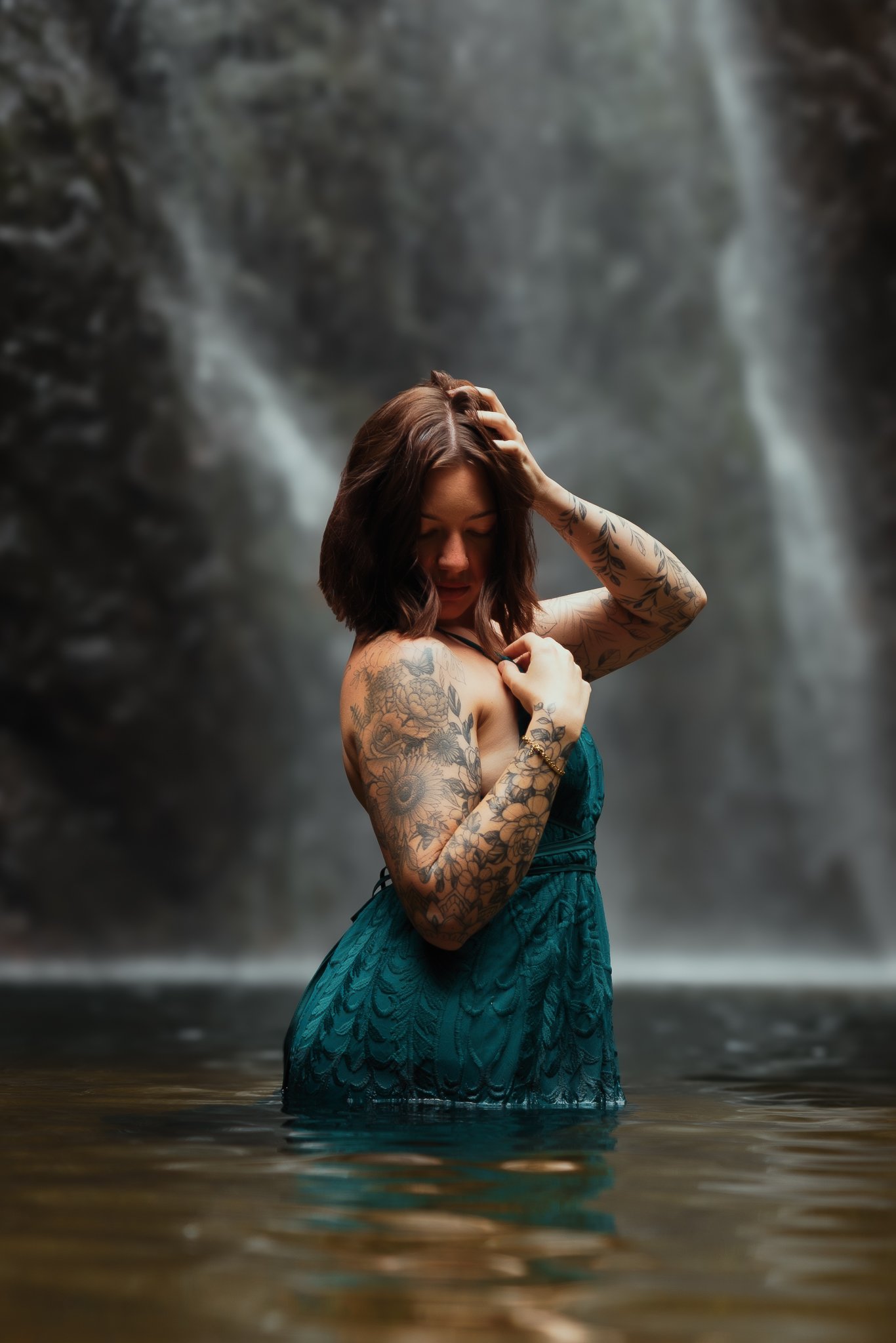 A woman with tattoos on her arms standing in water with a waterfall in the background, wearing a teal dress, with one hand on her head and the other near her chest.