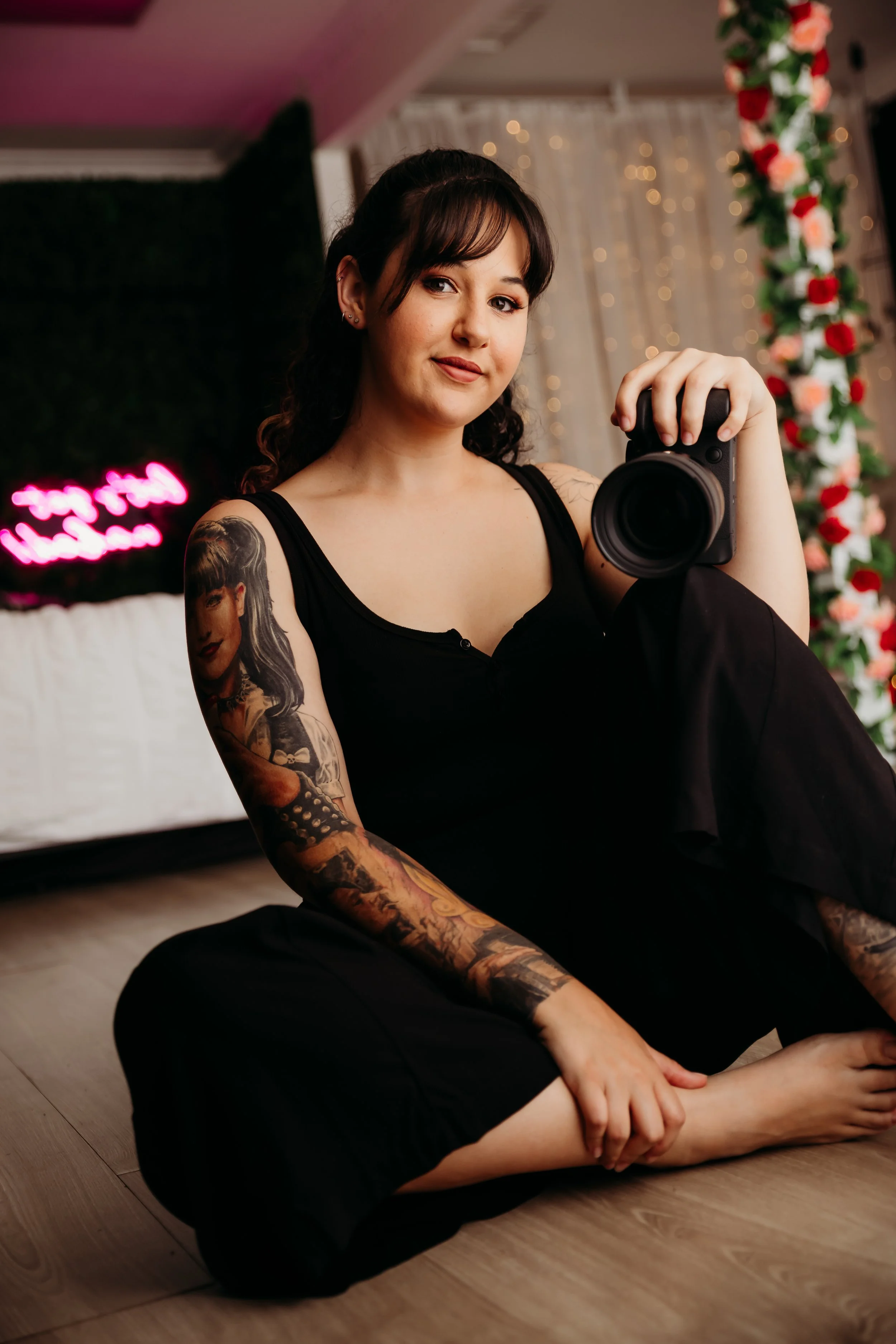 Pictured is Amy Davis - Business owner & Photographer at Temptations Intimate Photography. Young woman with tattoos sitting on the floor in a room with pink and clear fairy lights, holding a camera, and wearing a black sleeveless top and black pants.