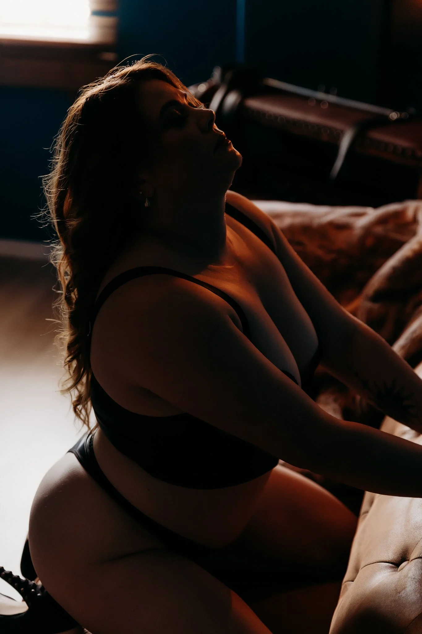 A woman with long hair and tattoos, in black lingerie, is kneeling on a couch with her eyes closed in a dimly lit room.