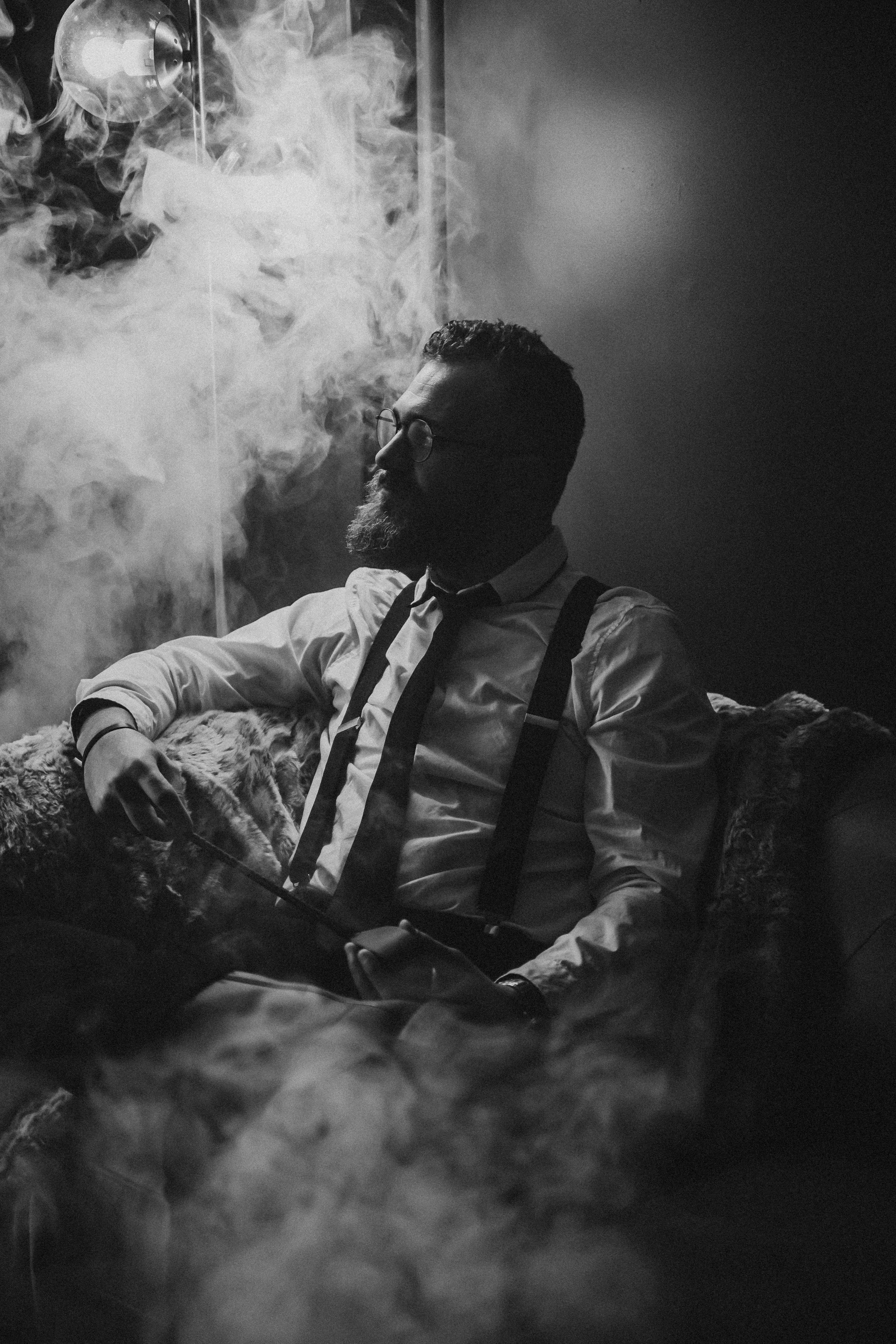 A black and white photo of a man with a beard and glasses, sitting on a couch with smoke around him, wearing a white shirt and suspenders, holding a cigarette.