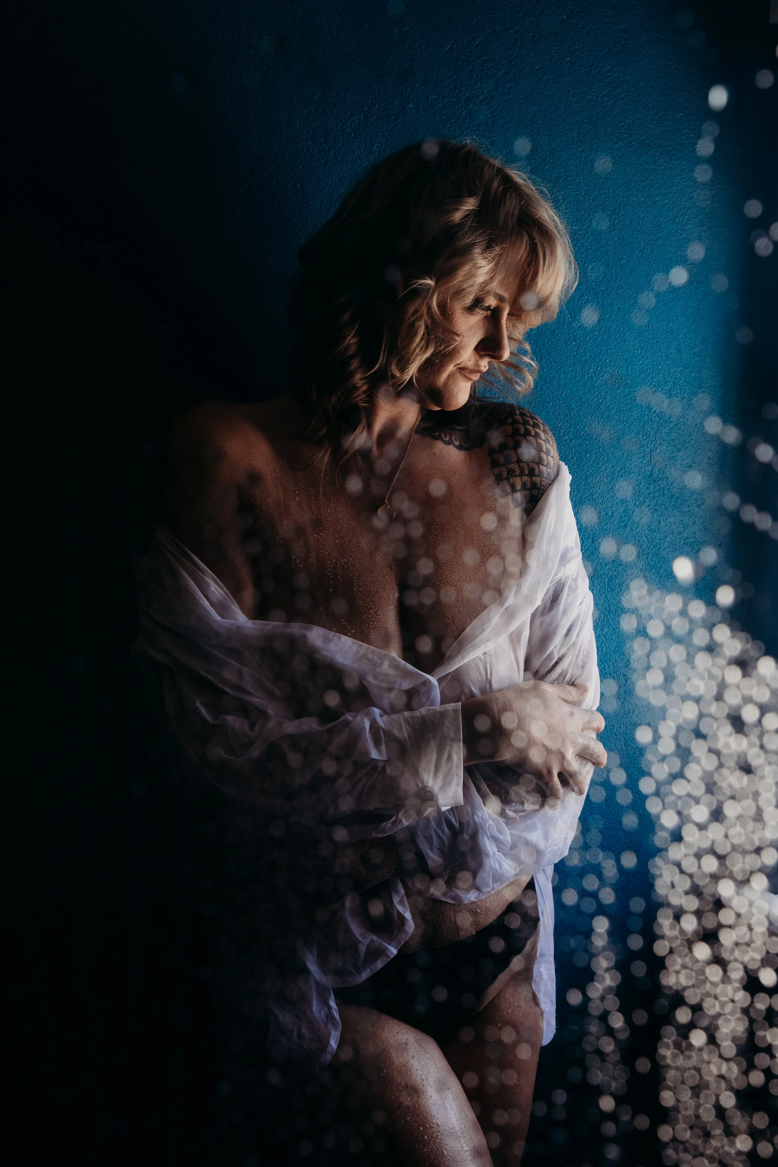 A woman with curly blonde hair standing against a blue wall, partially covered with a white shirt, looking down with a serene expression. Water droplets or mist are visible in the foreground and background.