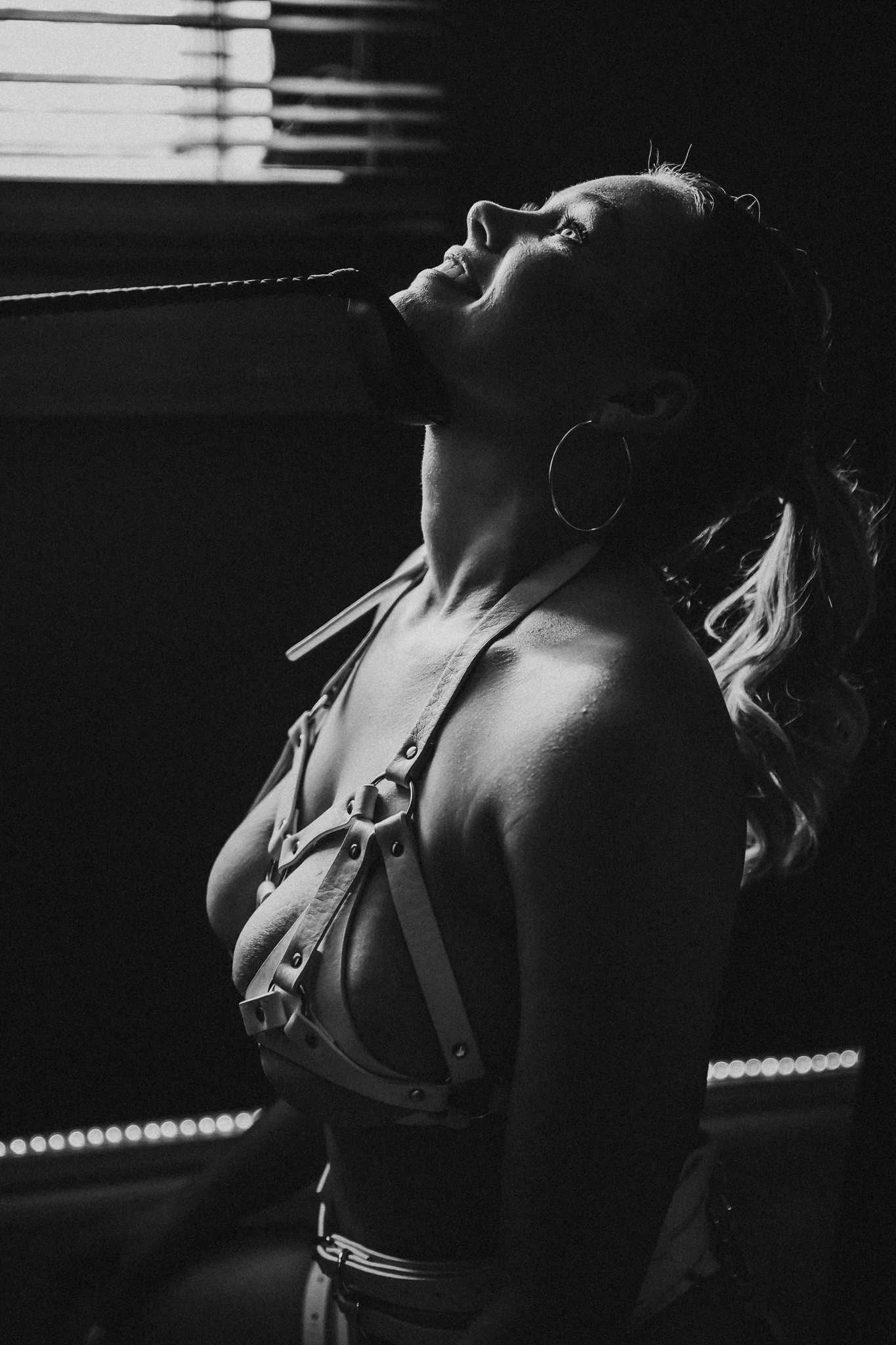 A woman wearing a harness poses in front of window blinds in a black and white photo.