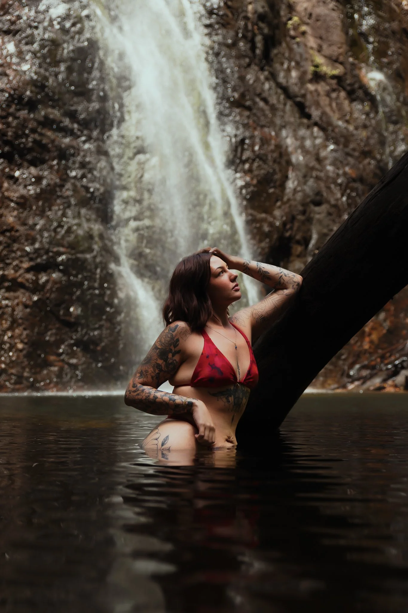 A woman with tattoos in a red bikini stands in water with a waterfall in the background, holding a log.