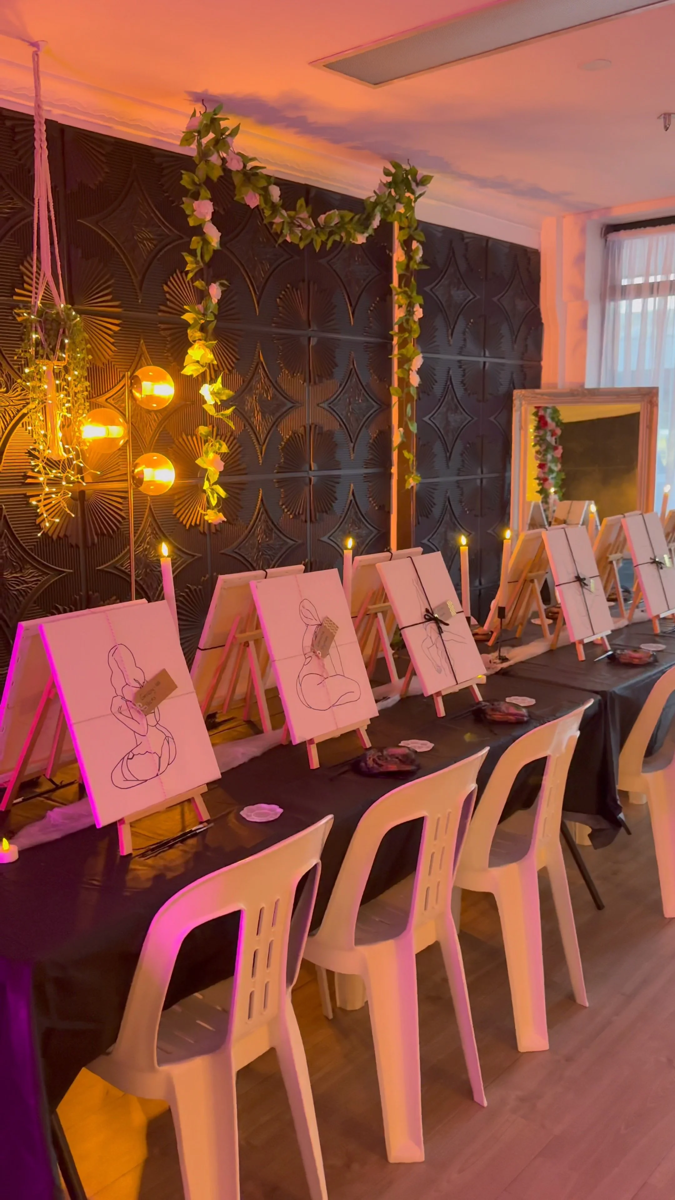 Indoor art setup with boudoir images outlined on canvases sitting on easels, lighting fixtures, and decorative greenery on a dark wall.