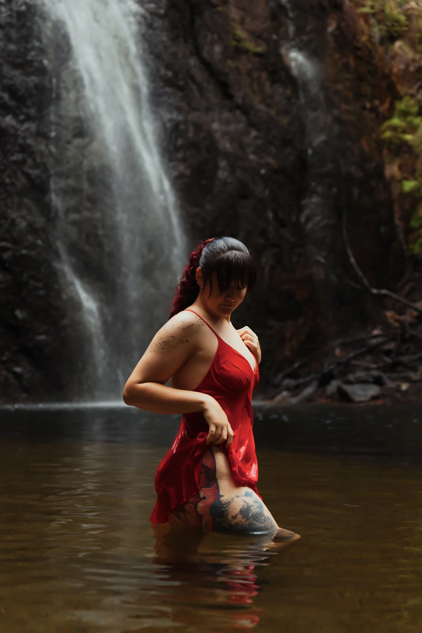 A woman wearing a red dress standing in a body of water with a waterfall in the background.