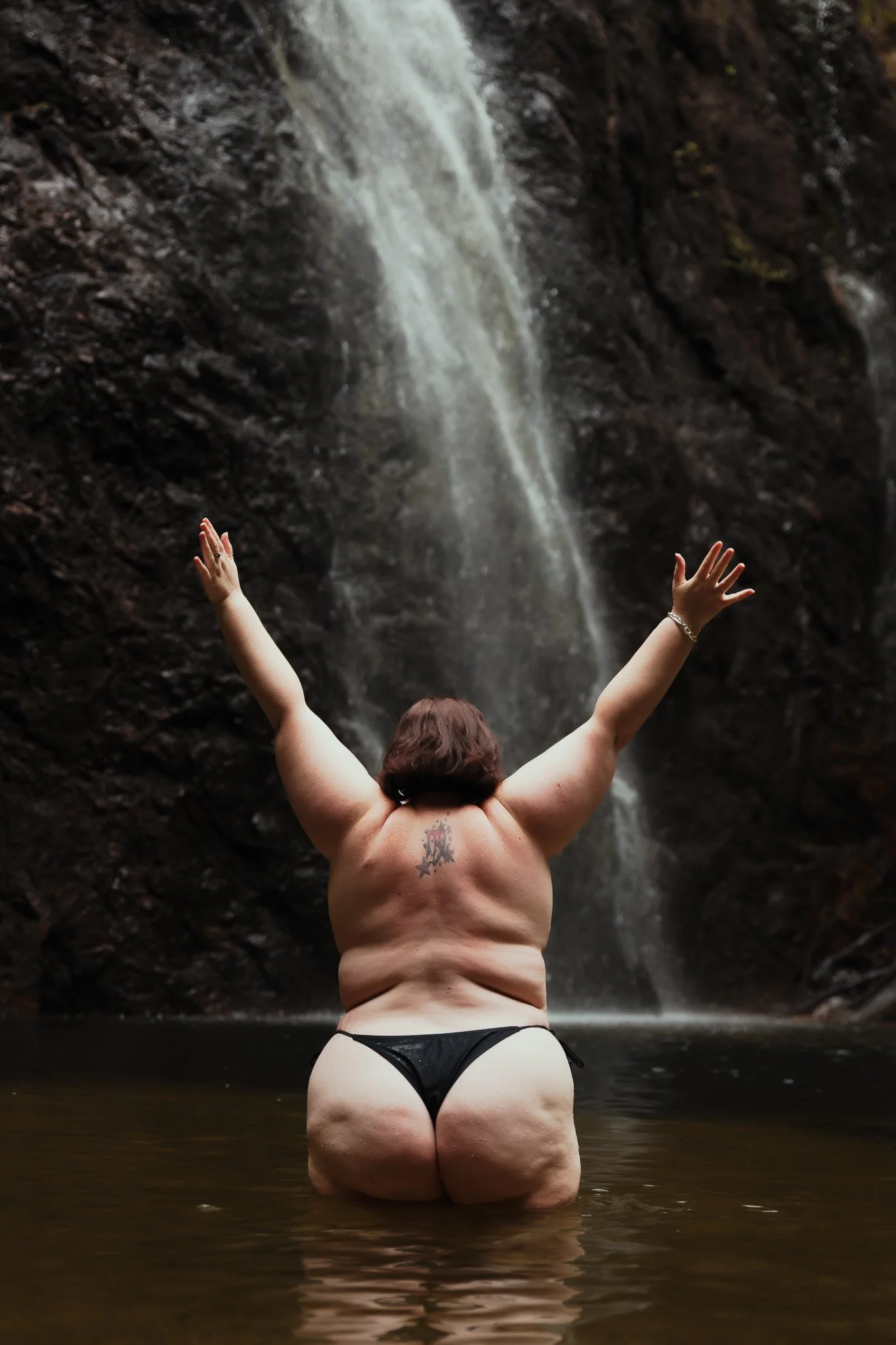 A person with tattoos and black swimwear standing in water with arms raised in front of a waterfall.
