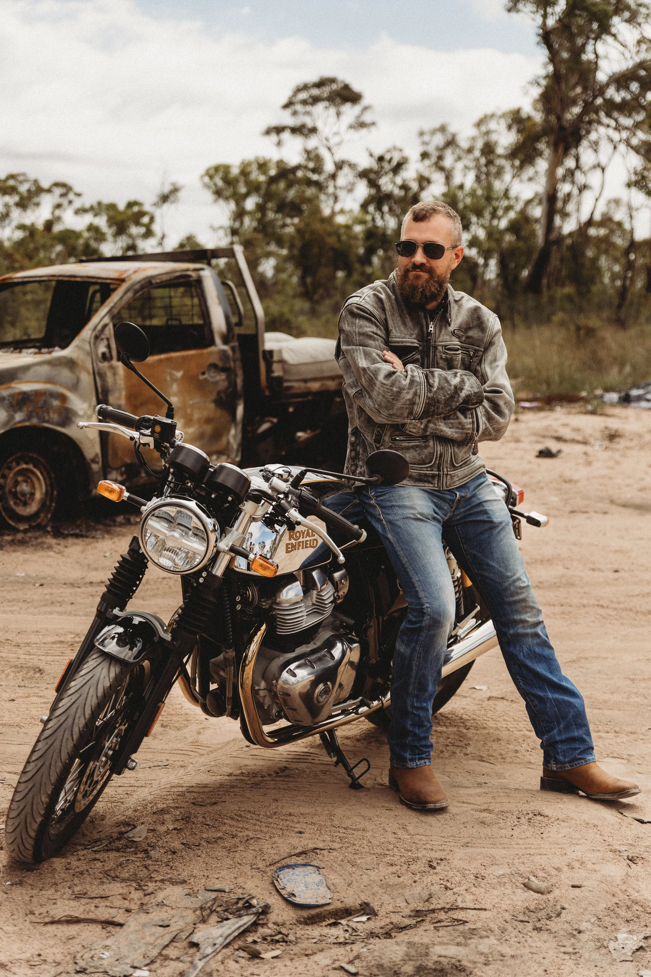 A man with sunglasses and a leather jacket sitting on a black and chrome Royal Enfield motorcycle in a dusty outdoor area, with a burned-out, rusted car in the background and trees and clouds overhead.