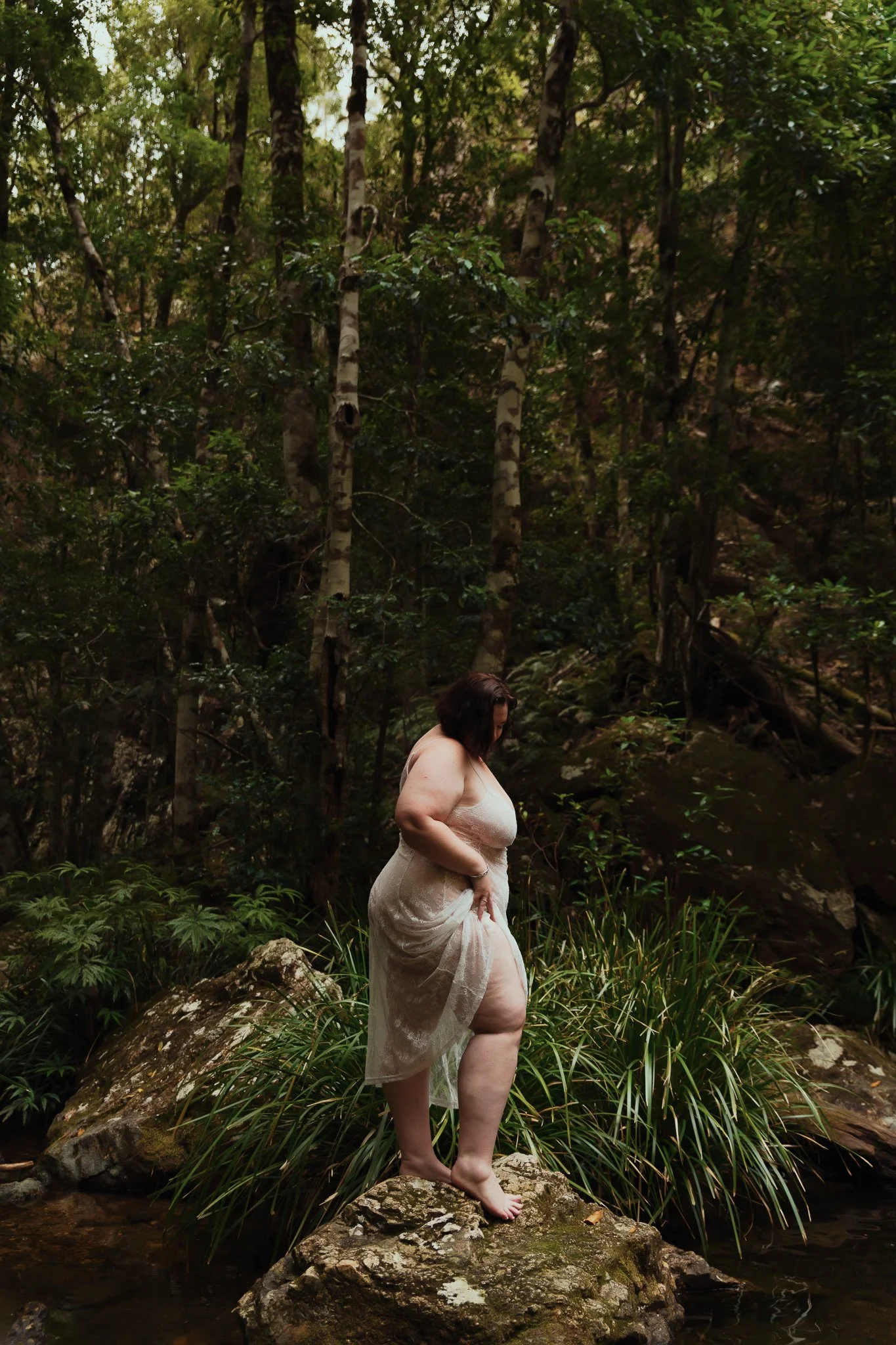 A woman in a semi-transparent light-colored dress standing barefoot on a rock in a forest with trees and green foliage.