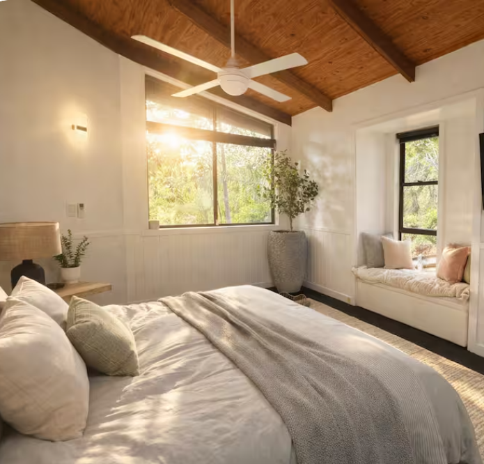 Bright bedroom with white walls, a bed with white bedding and pillows, a ceiling fan, large window with sunlight, a window seat with cushions, a potted plant, and wooden ceiling beams.