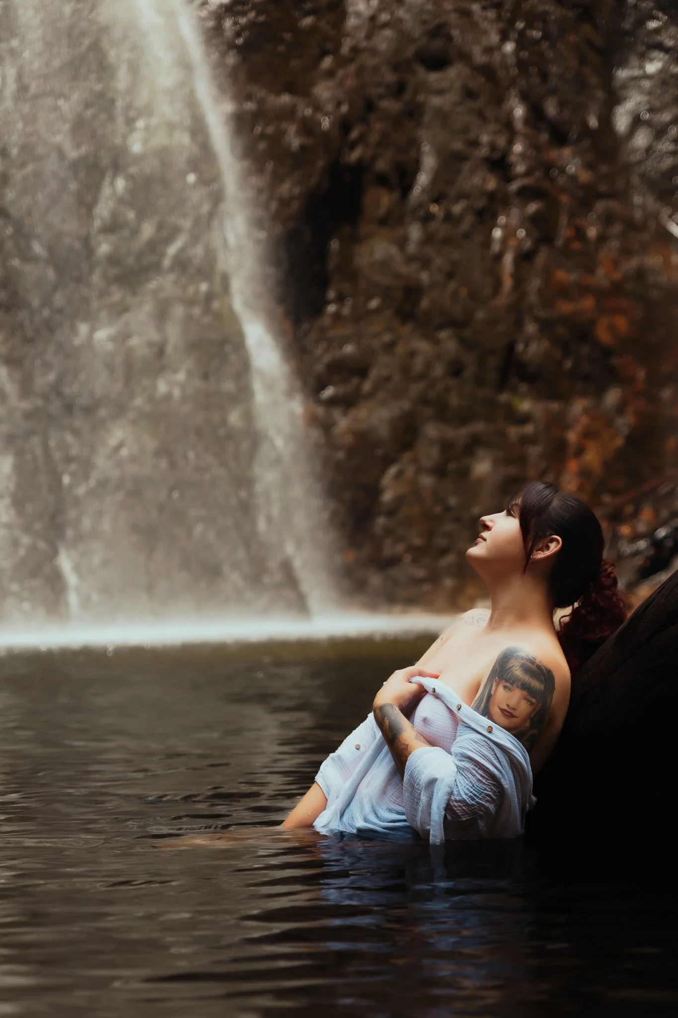 A woman with tattoos and dark hair, wearing a partially off-shoulder shirt, relaxing in water near a waterfall, with her eyes closed and face tilted upward.